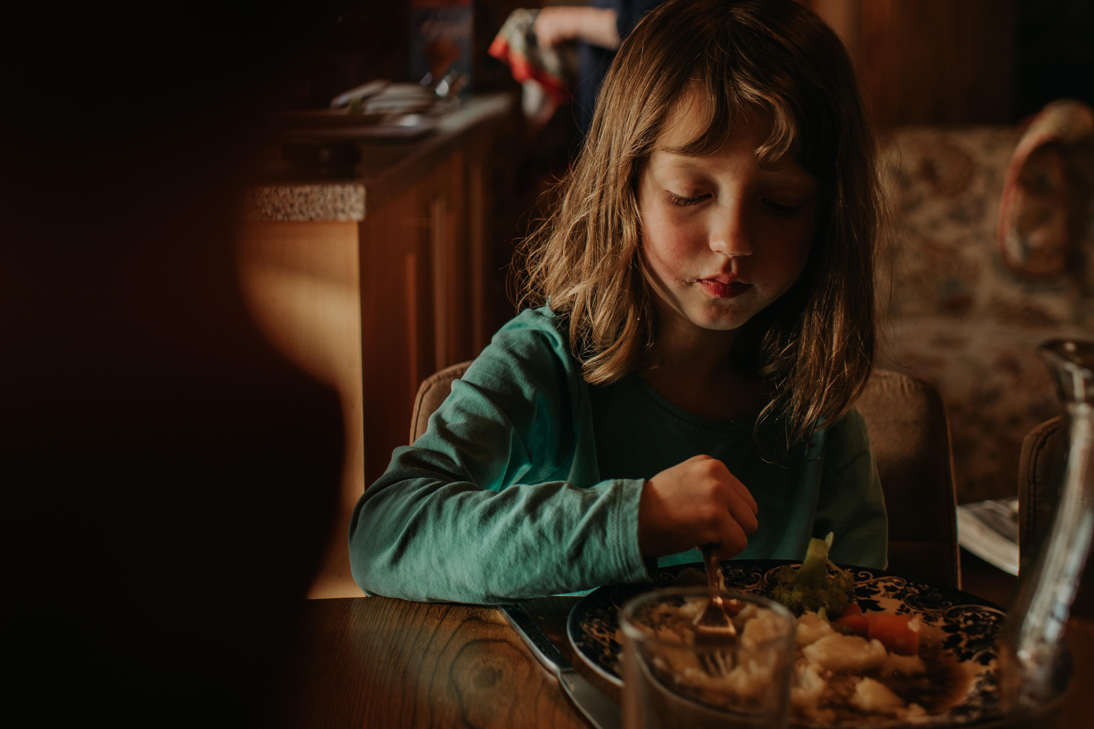 Photo of a young girl eating at a table indoors with an older man in the background, warmly lit setting.