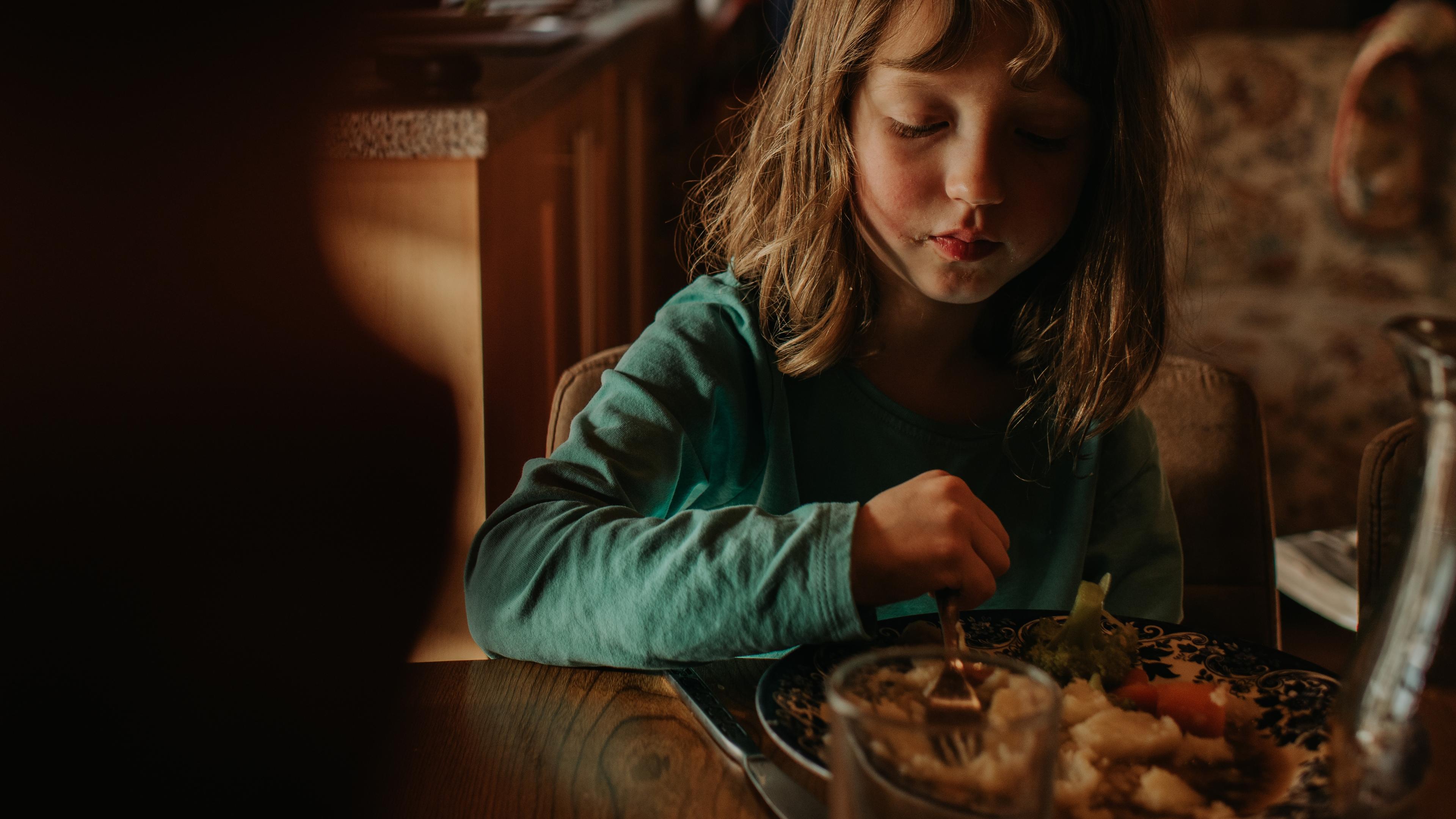 Photo of a young girl eating at a table indoors with an older man in the background, warmly lit setting.