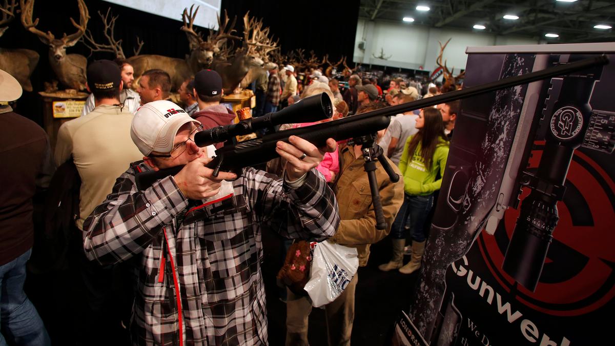 A man looking through the scope of a rifle in a crowded exhibition with mounted deer heads in the background.