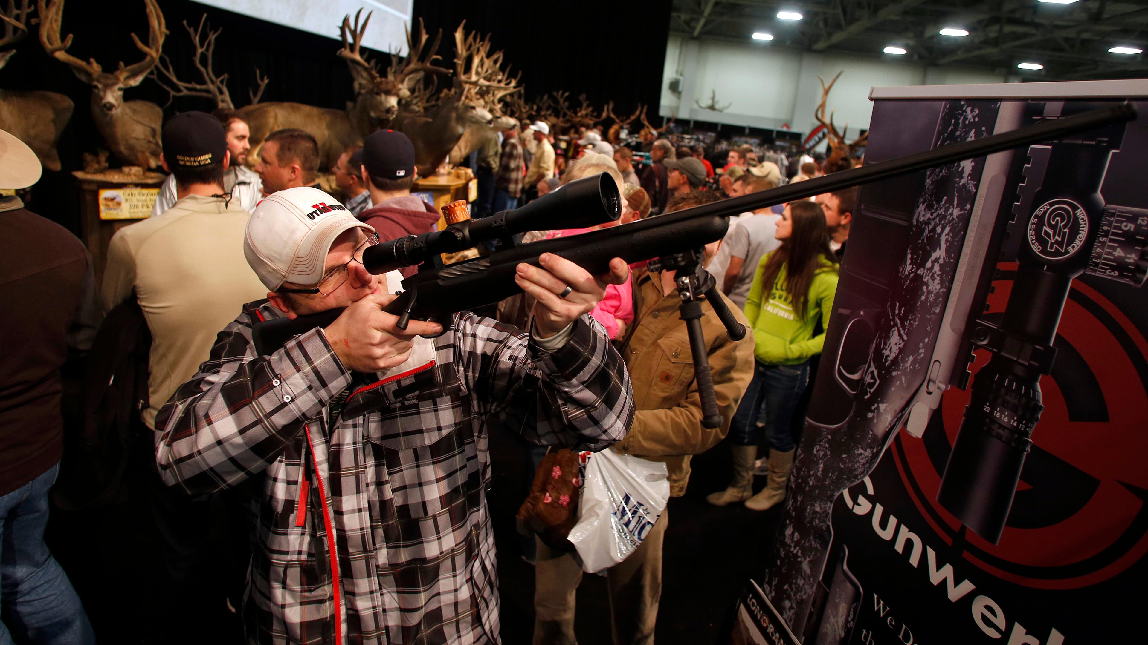 A man looking through the scope of a rifle in a crowded exhibition with mounted deer heads in the background.