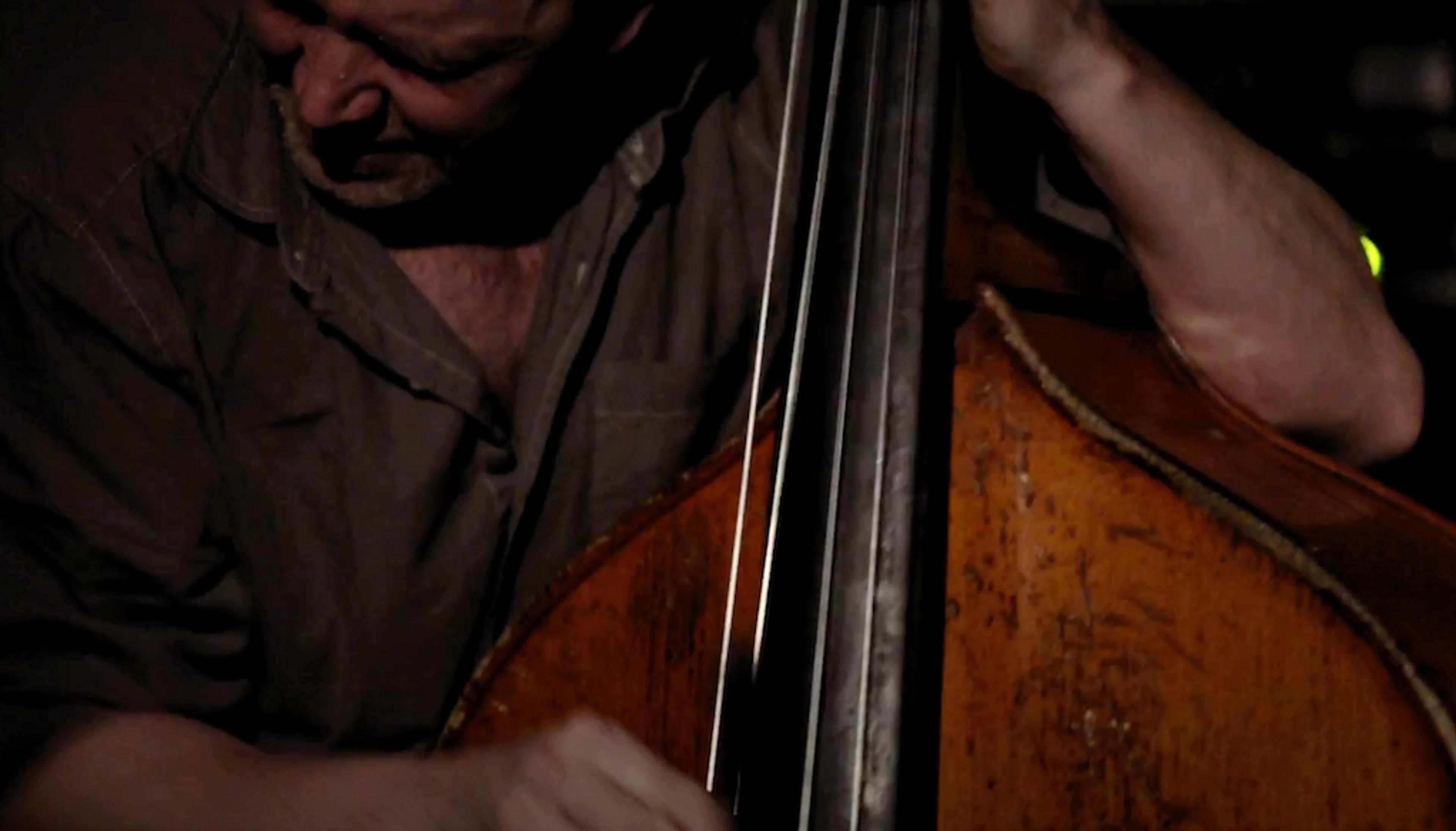 A musician intensely playing an upright bass, the focus is on his hands and the instrument’s worn, wooden surface.