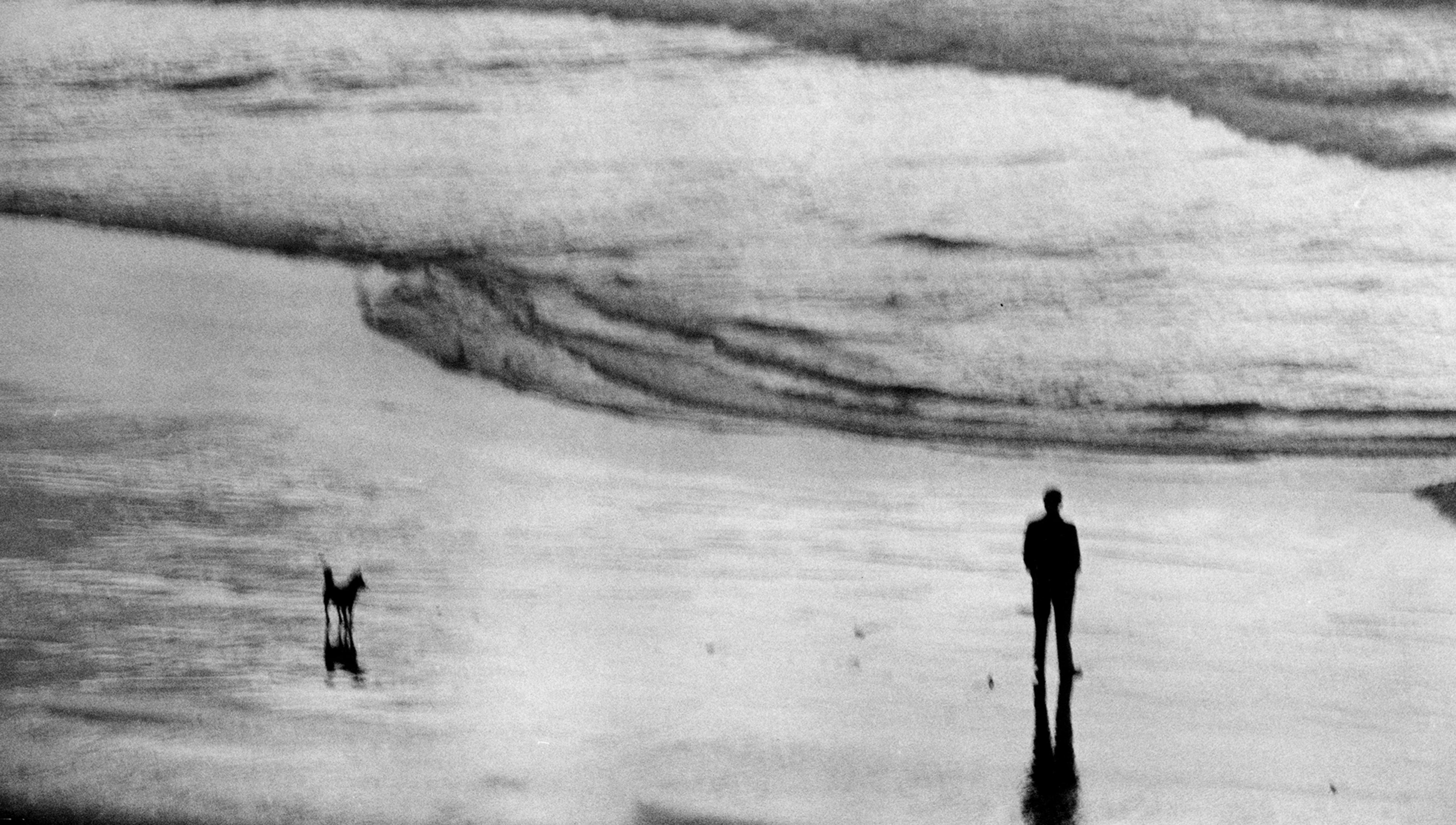 A monochrome photograph from above of a man looking out to sea. His hands are in his pockets and a dog waits some metres away