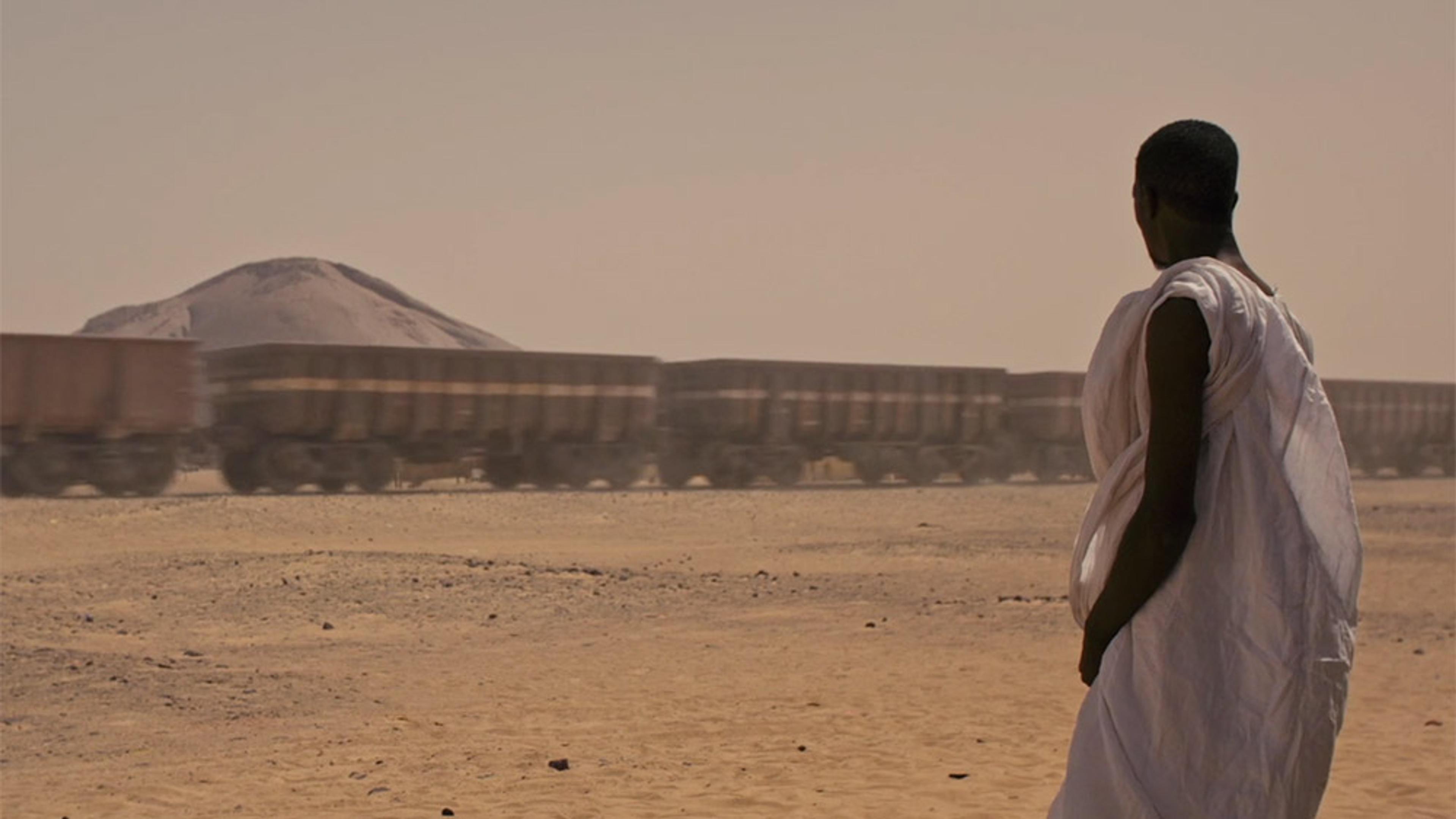 A person in traditional white attire watching a moving freight train in a desert landscape with a distant hill.