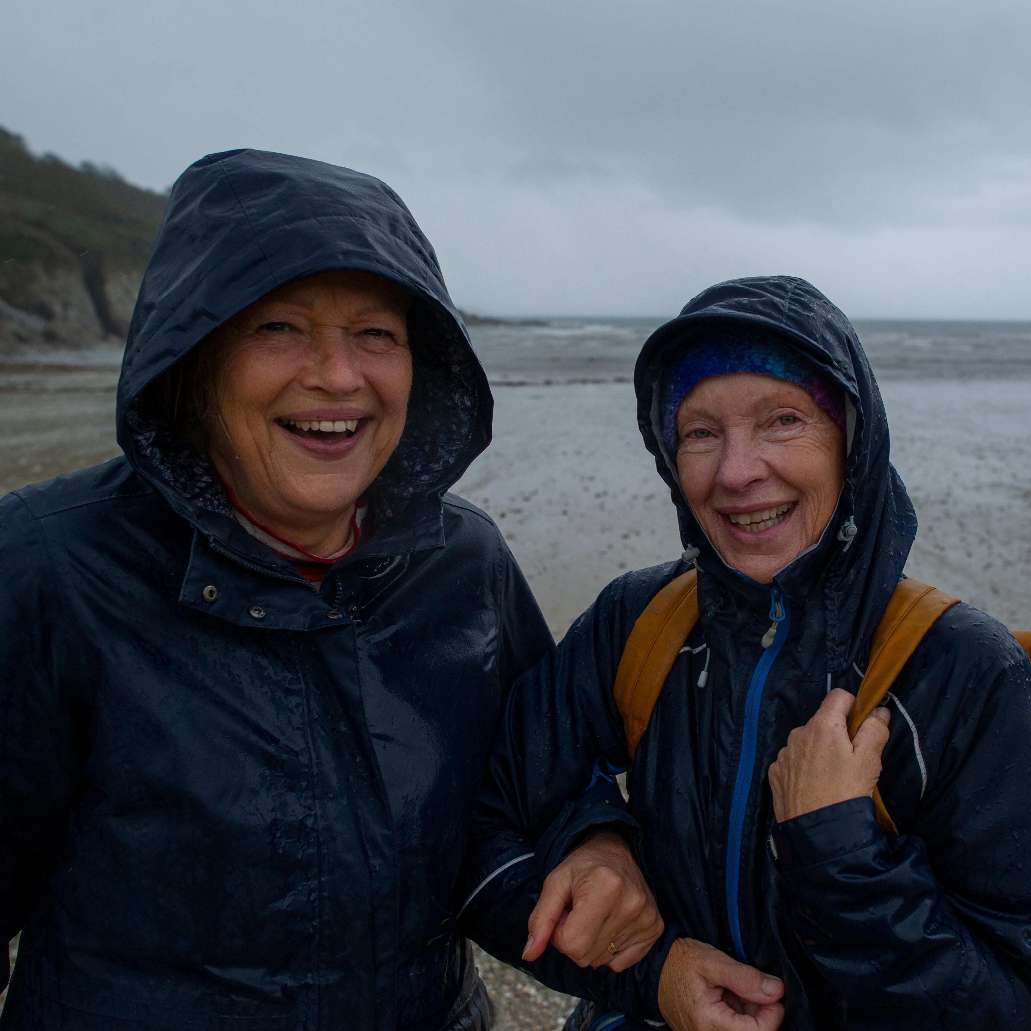 Photo of two smiling people in waterproof jackets on a rainy beach, grey skies in the background.