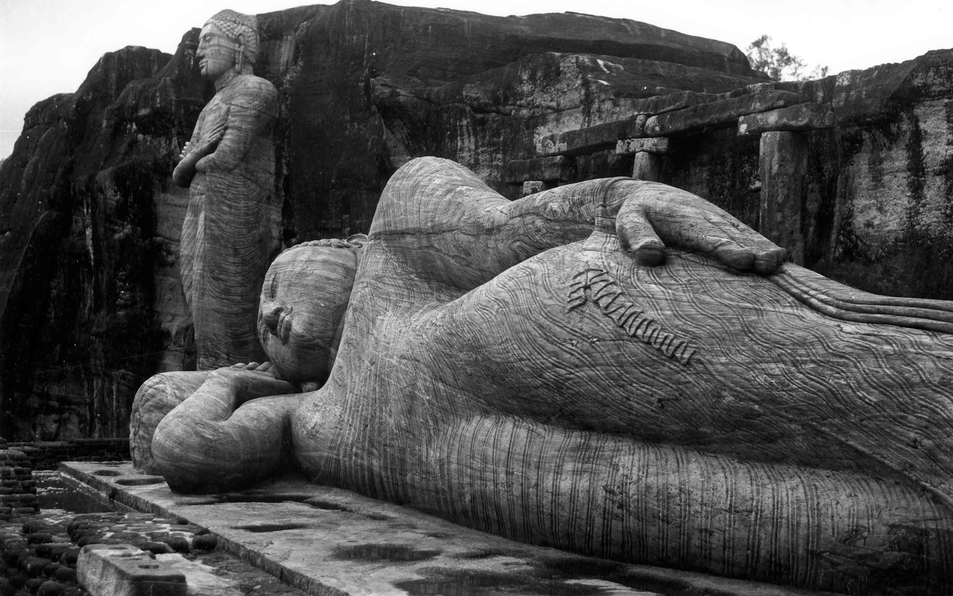 Ancient stone statues depicting a standing and a reclining Buddha against a natural rock backdrop.