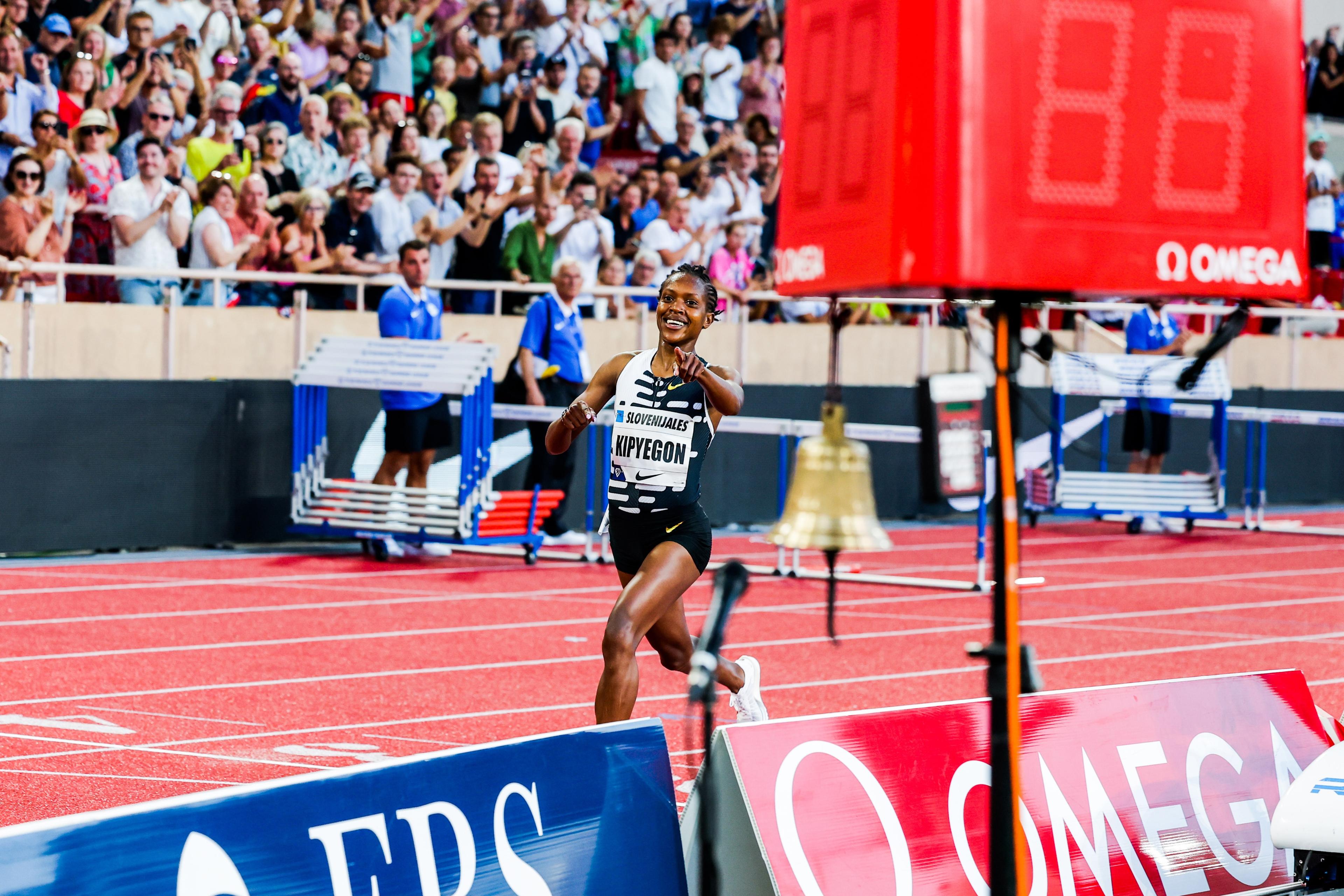 Photo of a female athlete smiling while crossing a track finish line with a cheering crowd in the background.