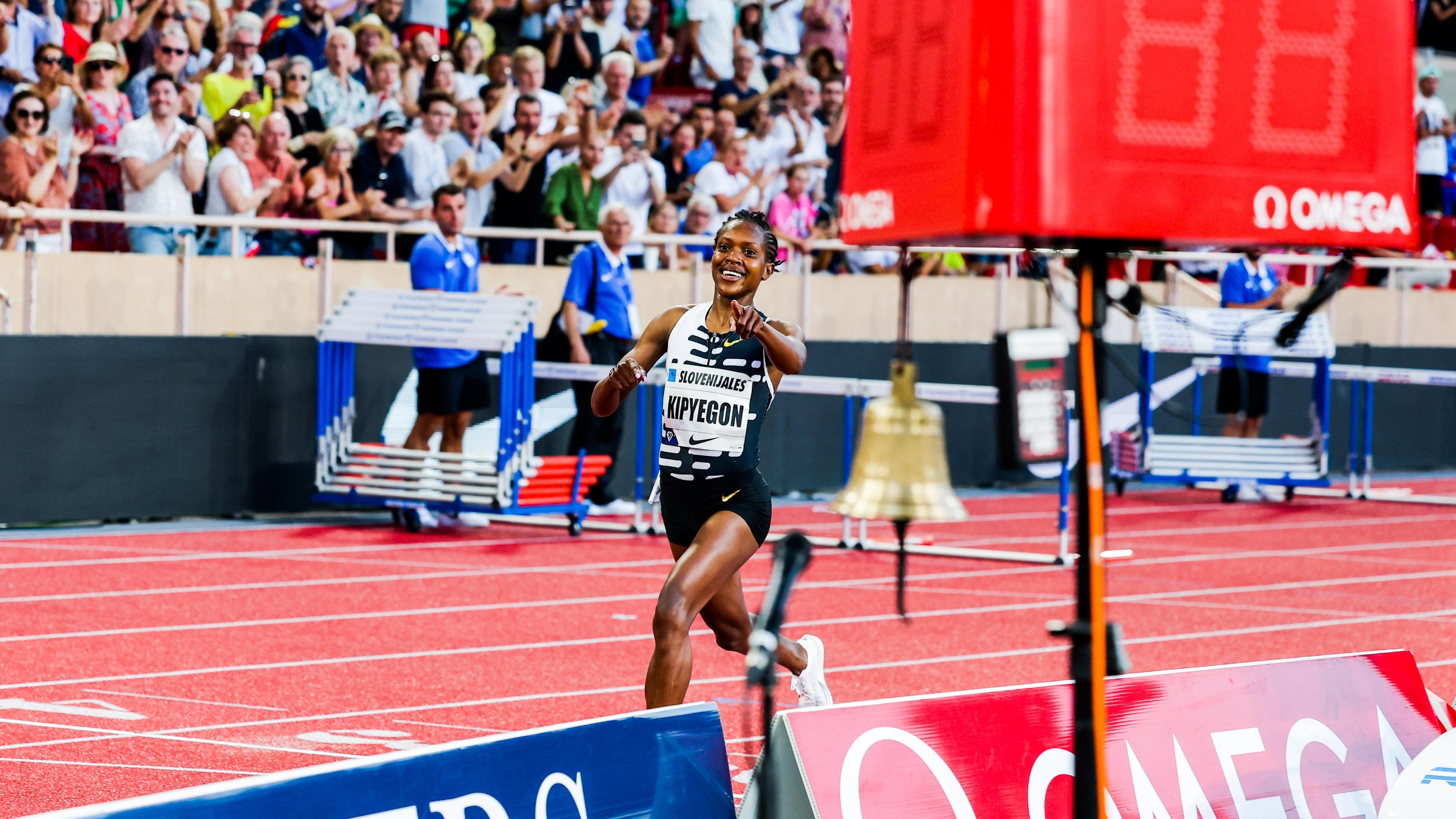 Photo of a female athlete smiling while crossing a track finish line with a cheering crowd in the background.