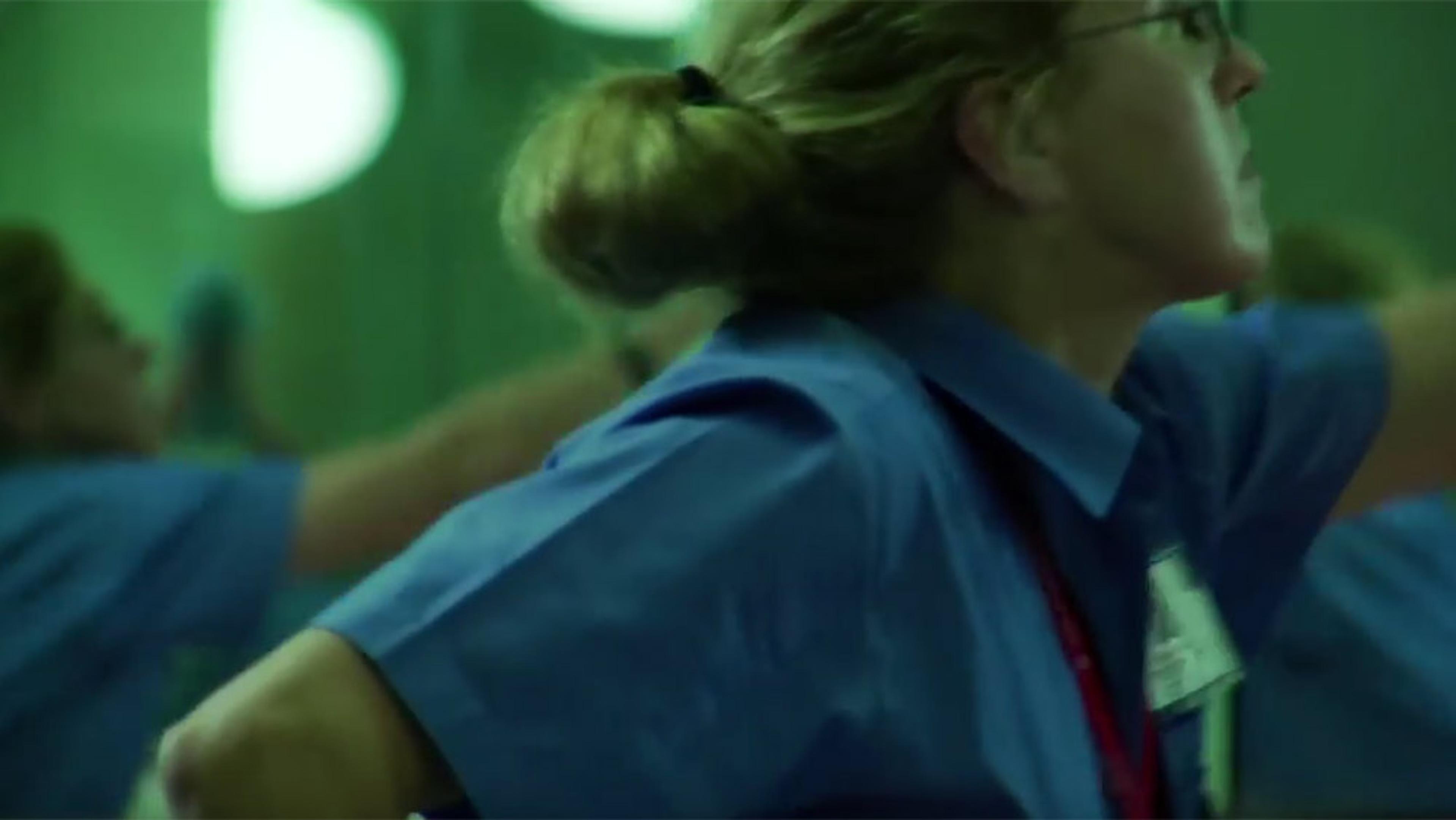 A woman in a blue uniform with a name tag, reaching out her arm with an intense expression, green background.
