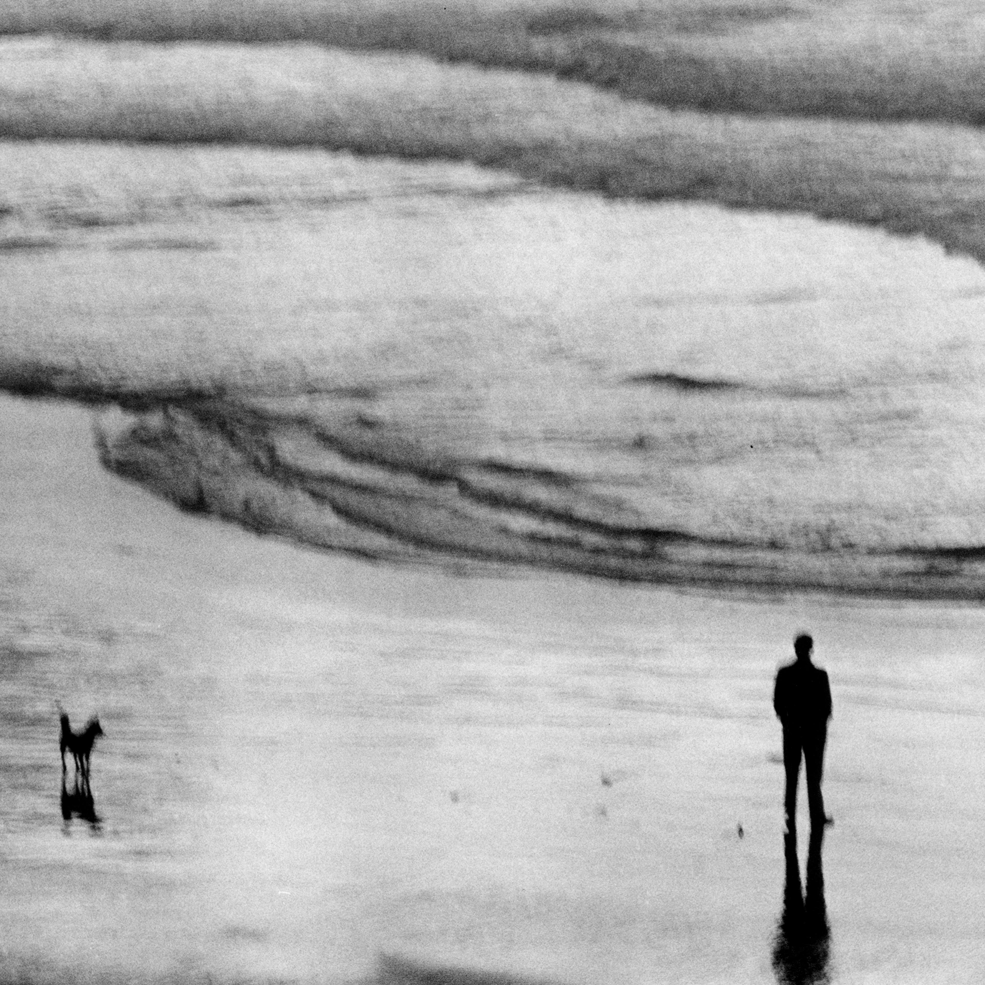 A monochrome photograph from above of a man looking out to sea. His hands are in his pockets and a dog waits some metres away