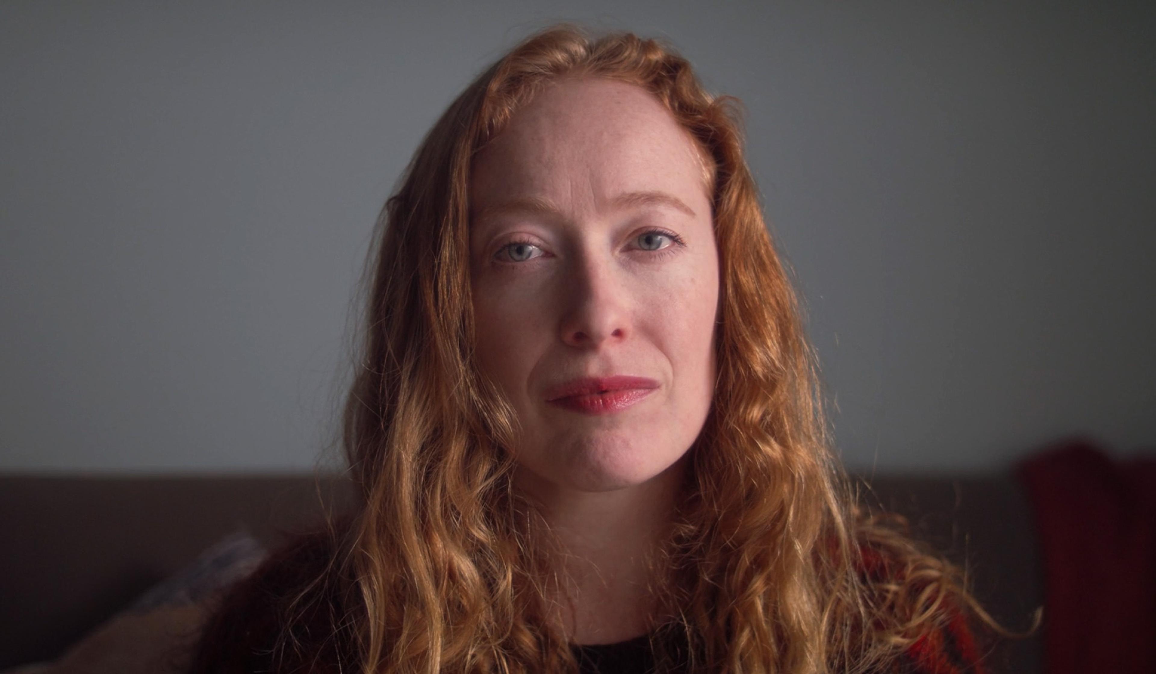 A woman with long, wavy red hair and a neutral expression, seated indoors with a blurred background.