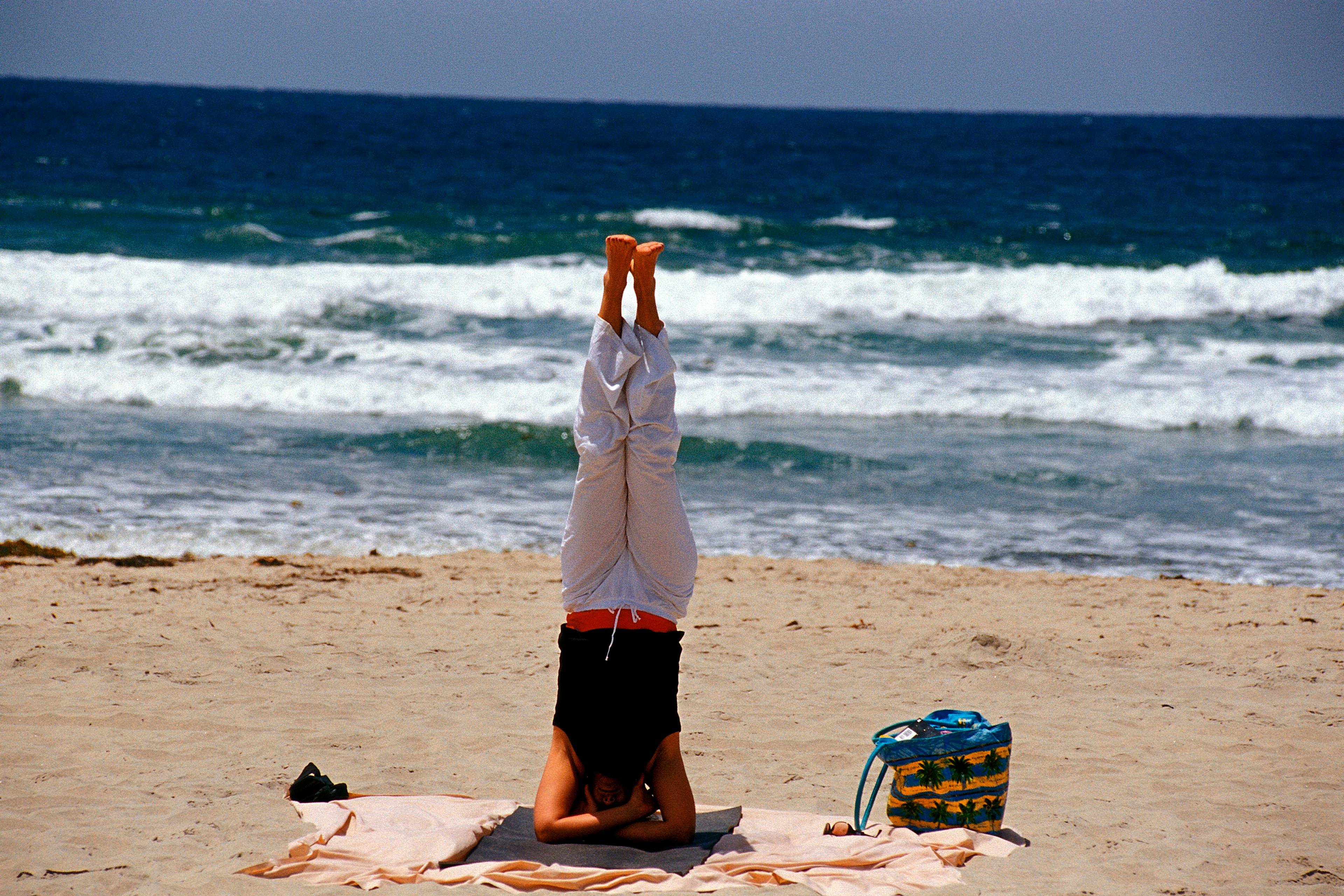 Photo of a person doing a headstand on a beach towel by the ocean, with a bag nearby and waves in the background.