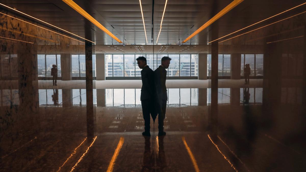 A man in silhouette standing against a reflective surface in an empty room with cityscape visible through large windows.