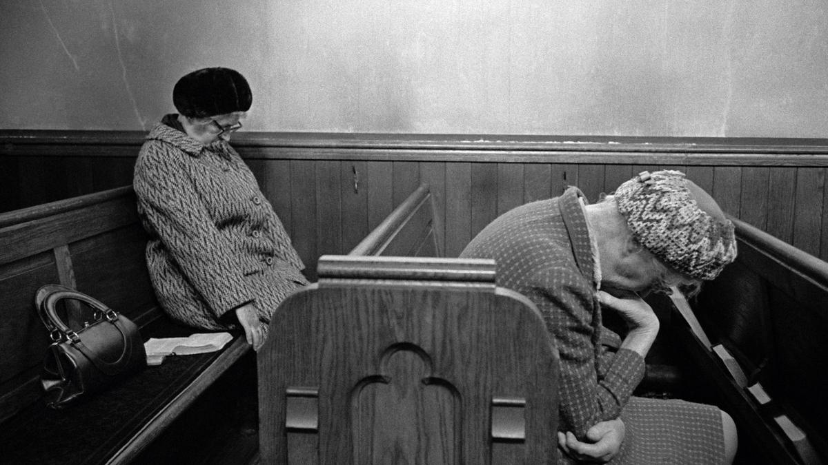 Black and white photo of two elderly women sitting on a wooden bench in a chapel with heads bowed, one has a handbag beside her.