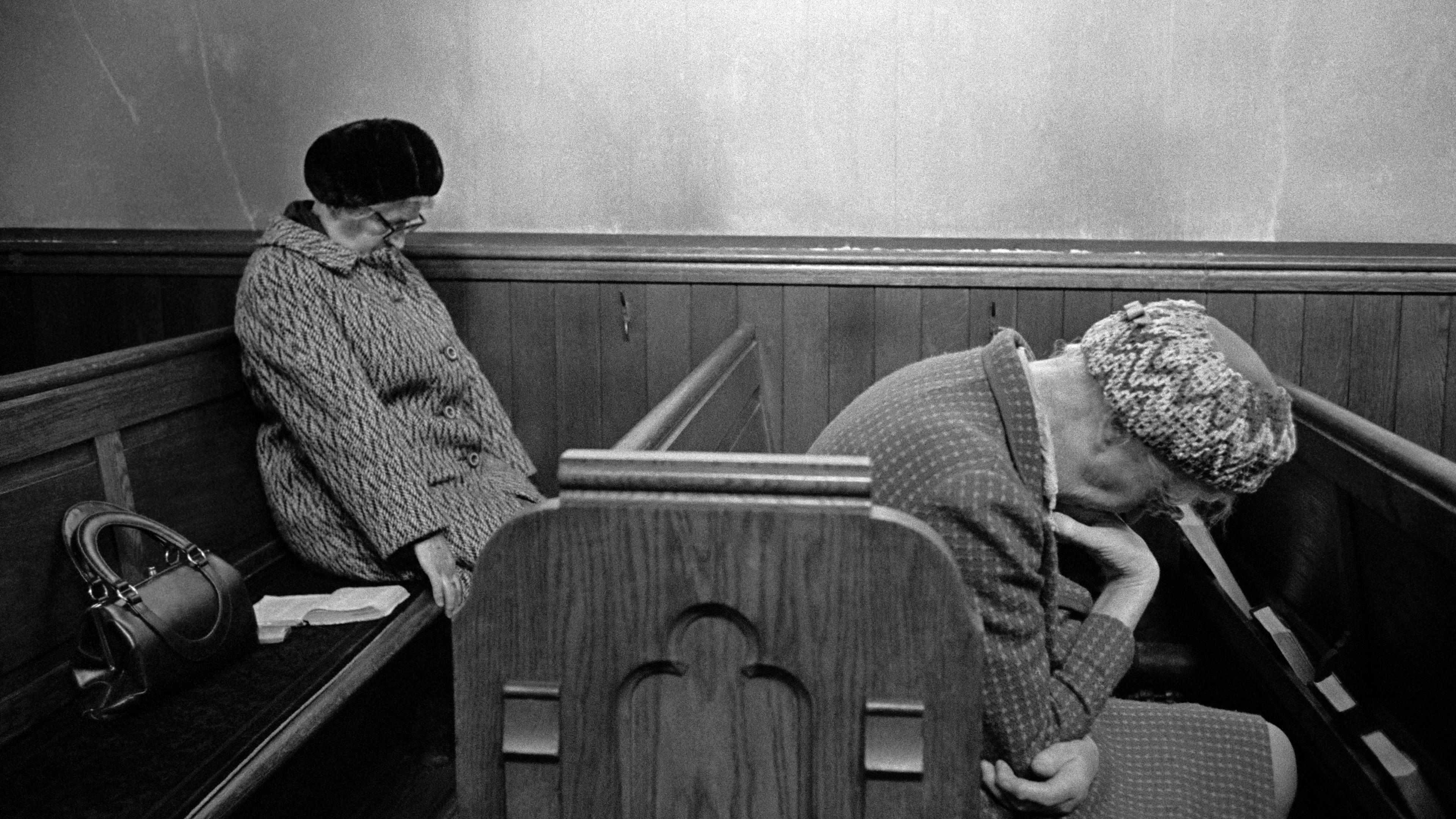 Black and white photo of two elderly women sitting on a wooden bench in a chapel with heads bowed, one has a handbag beside her.