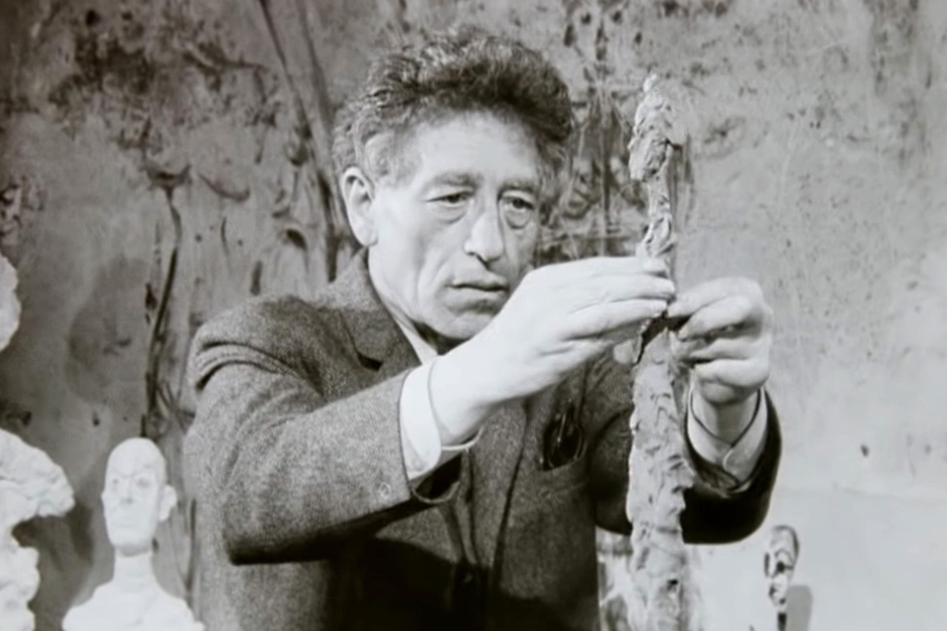 Black-and-white photo of a man in a suit who is Giacometti sculpting a thin figure, surrounded by small sculpted busts, in a studio with textured walls.