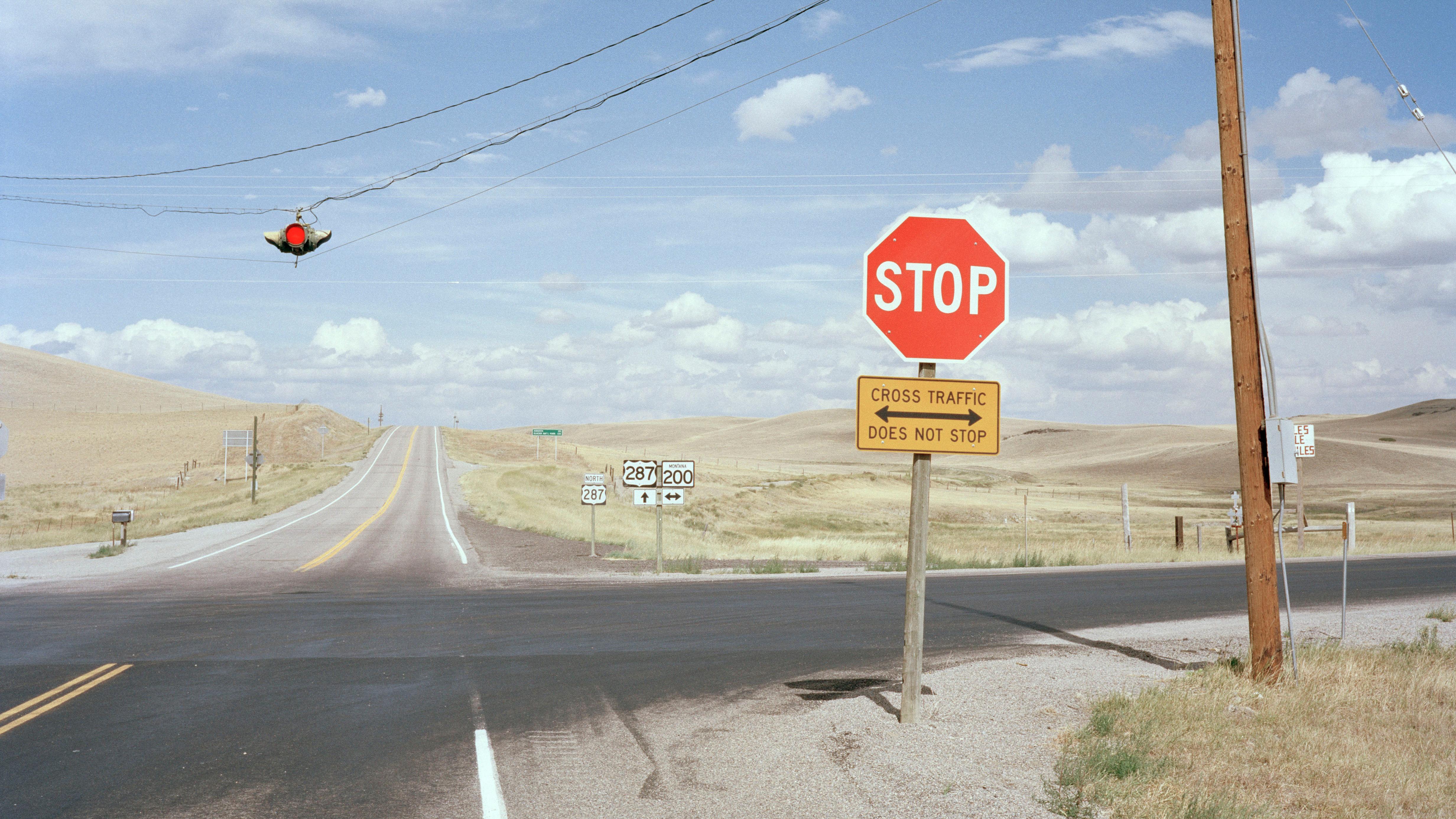 Photo of a rural crossroads with a stop sign, traffic lights above, a utility pole and vast open fields under a blue sky.