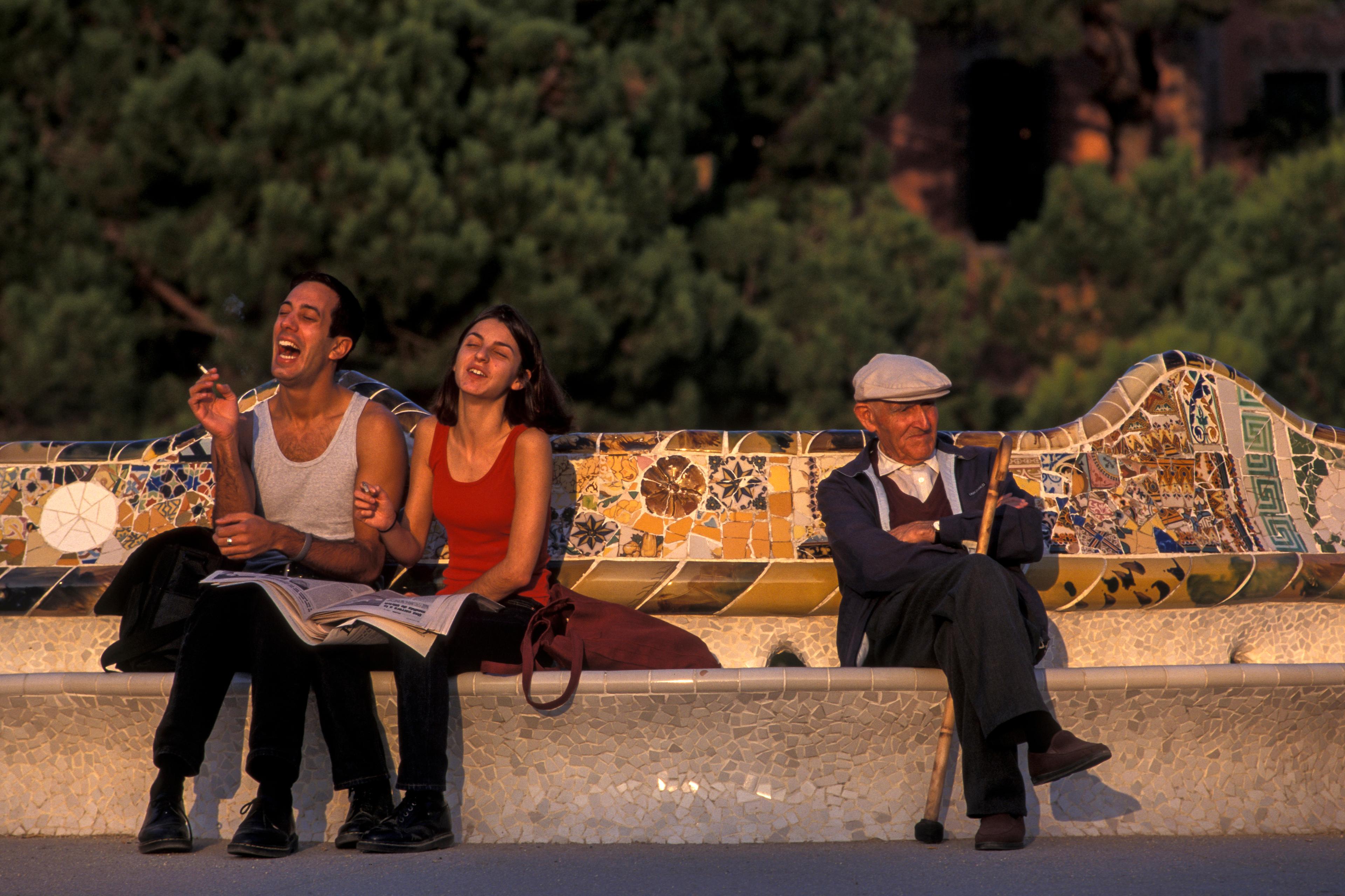 Photo of two people laughing and an elderly man sitting on a mosaic bench in a park with green trees in the background.