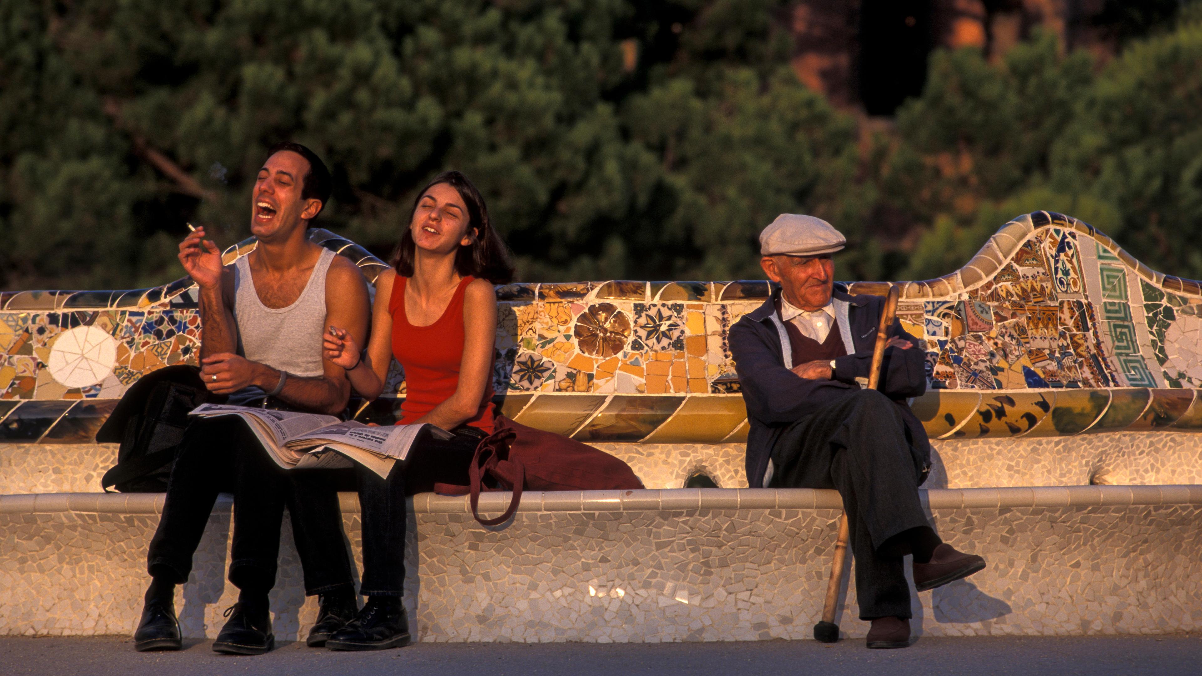 Photo of two people laughing and an elderly man sitting on a mosaic bench in a park with green trees in the background.