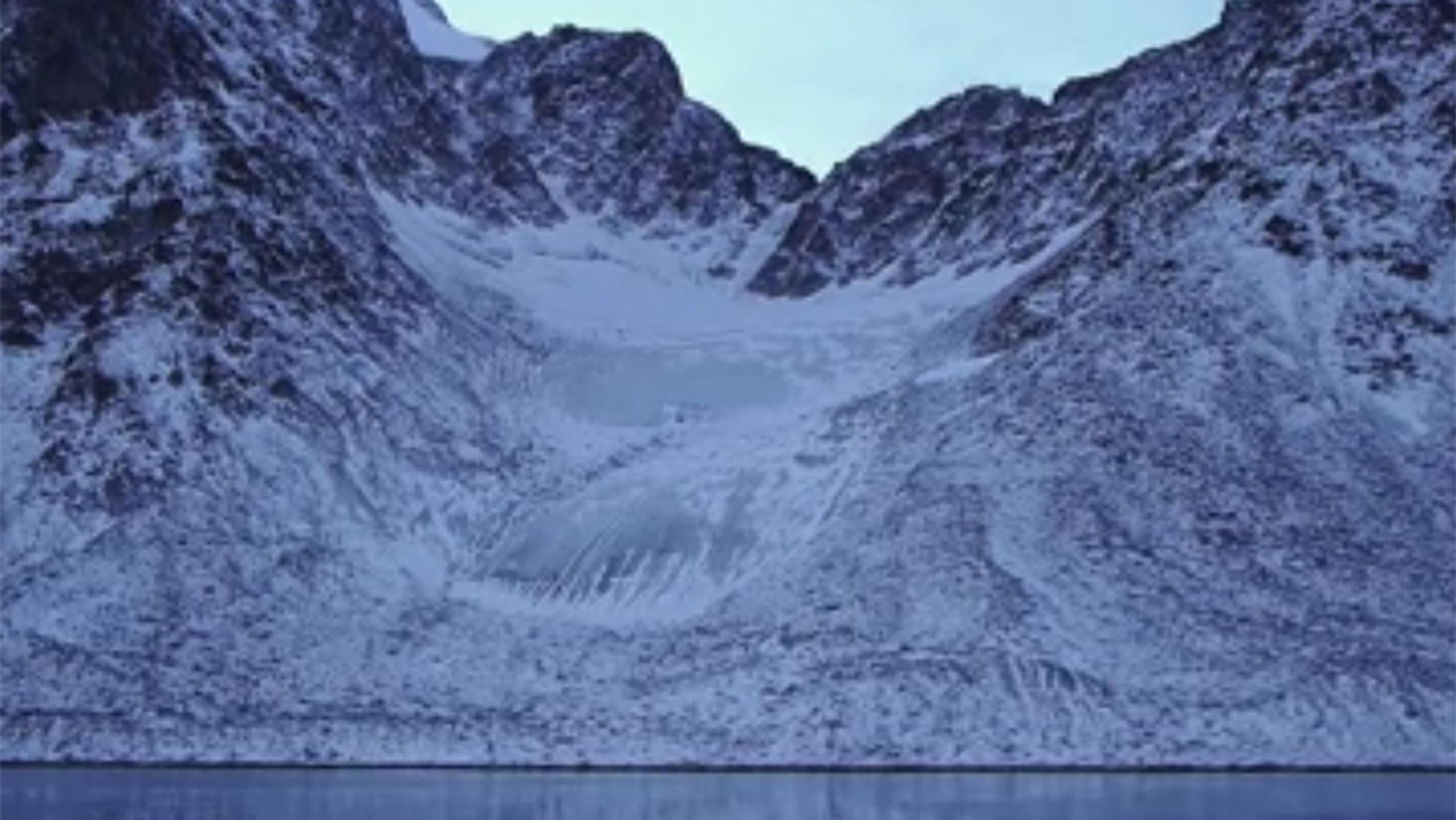 A snow-covered mountain range with a glacier in the centre, reflecting on a calm body of water below.