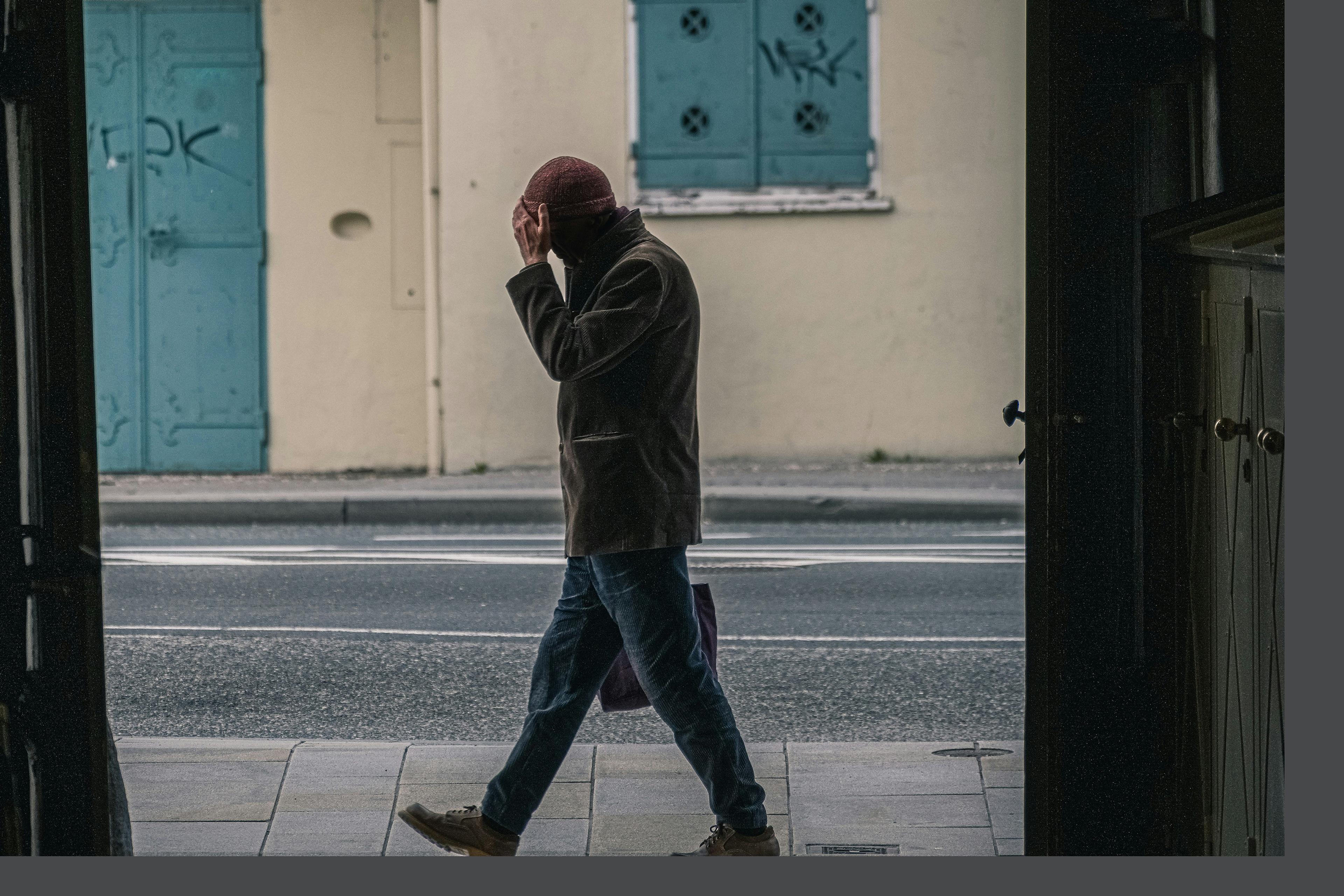 A person walking on a pavement, holding their head, with blue shutters and a lightly graffitied wall in the background.