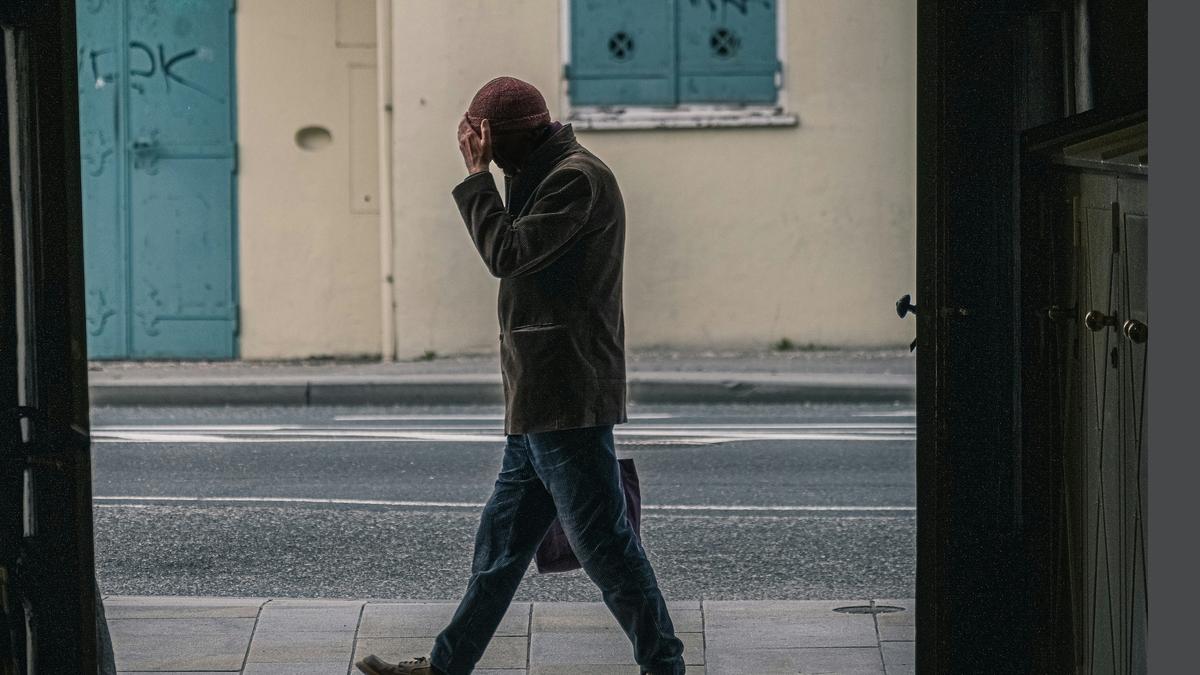 A person walking on a pavement, holding their head, with blue shutters and a lightly graffitied wall in the background.