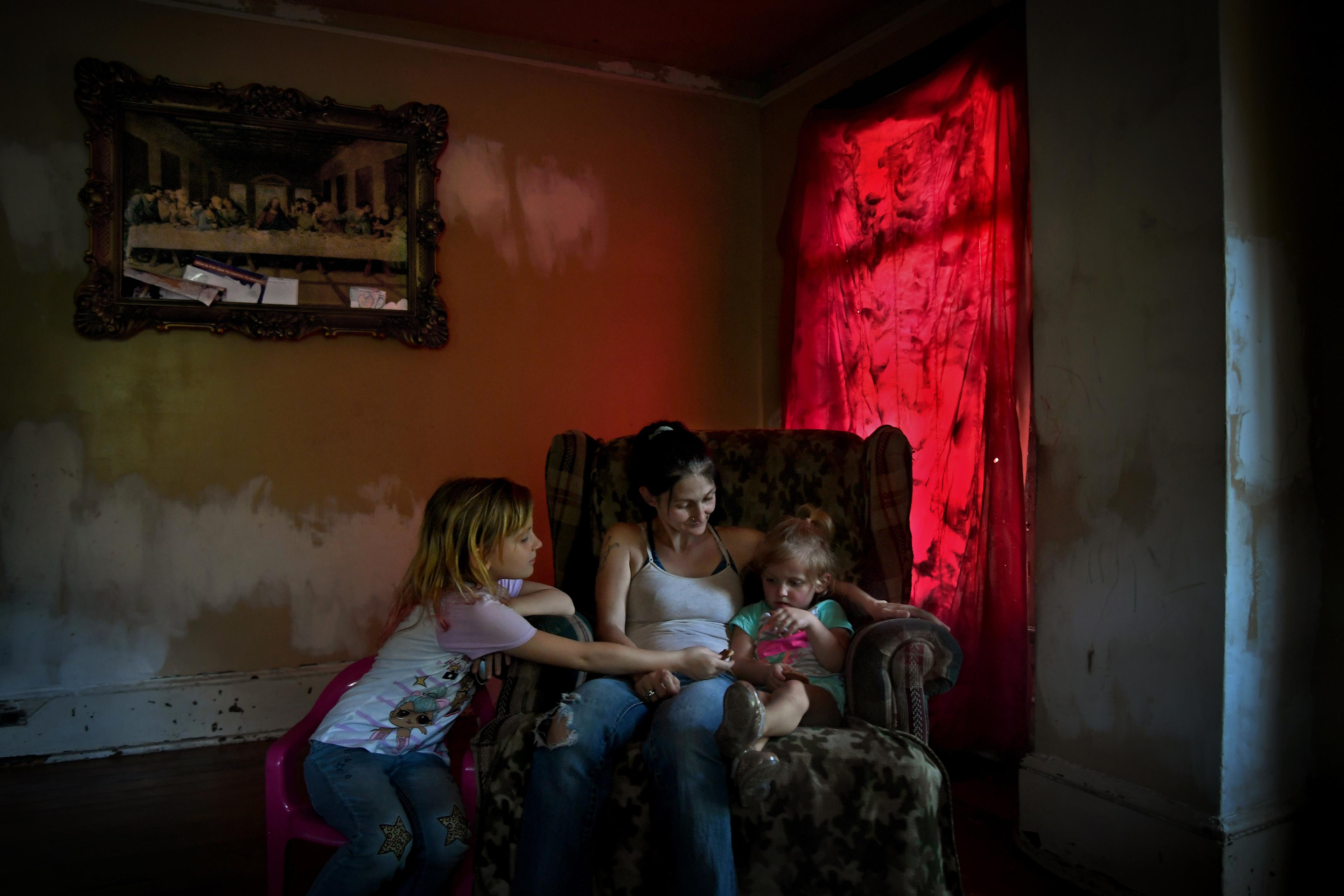Photo of a woman and two children sitting in an armchair in a dimly lit room with red curtains and a framed painting.