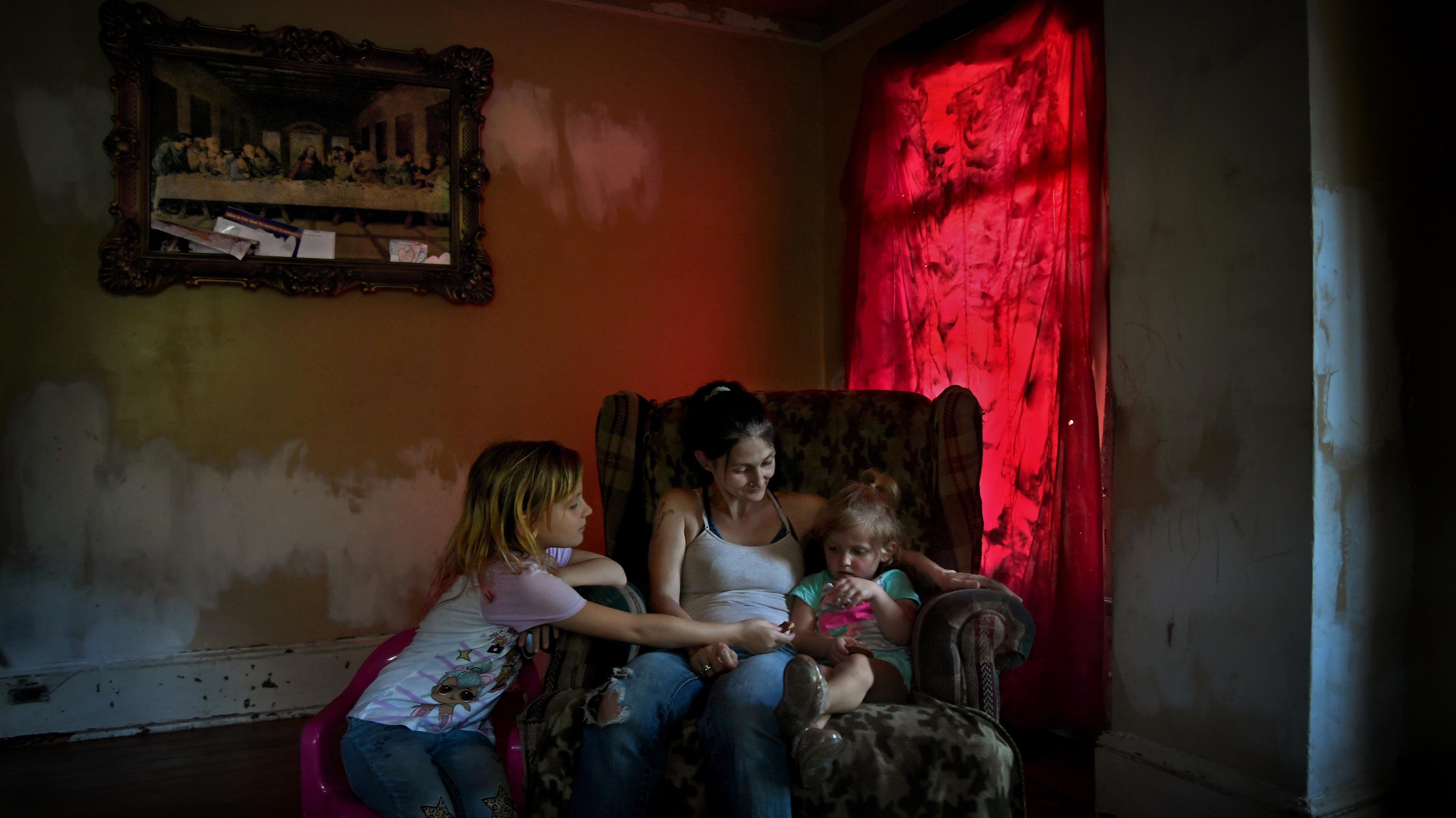 Photo of a woman and two children sitting in an armchair in a dimly lit room with red curtains and a framed painting.