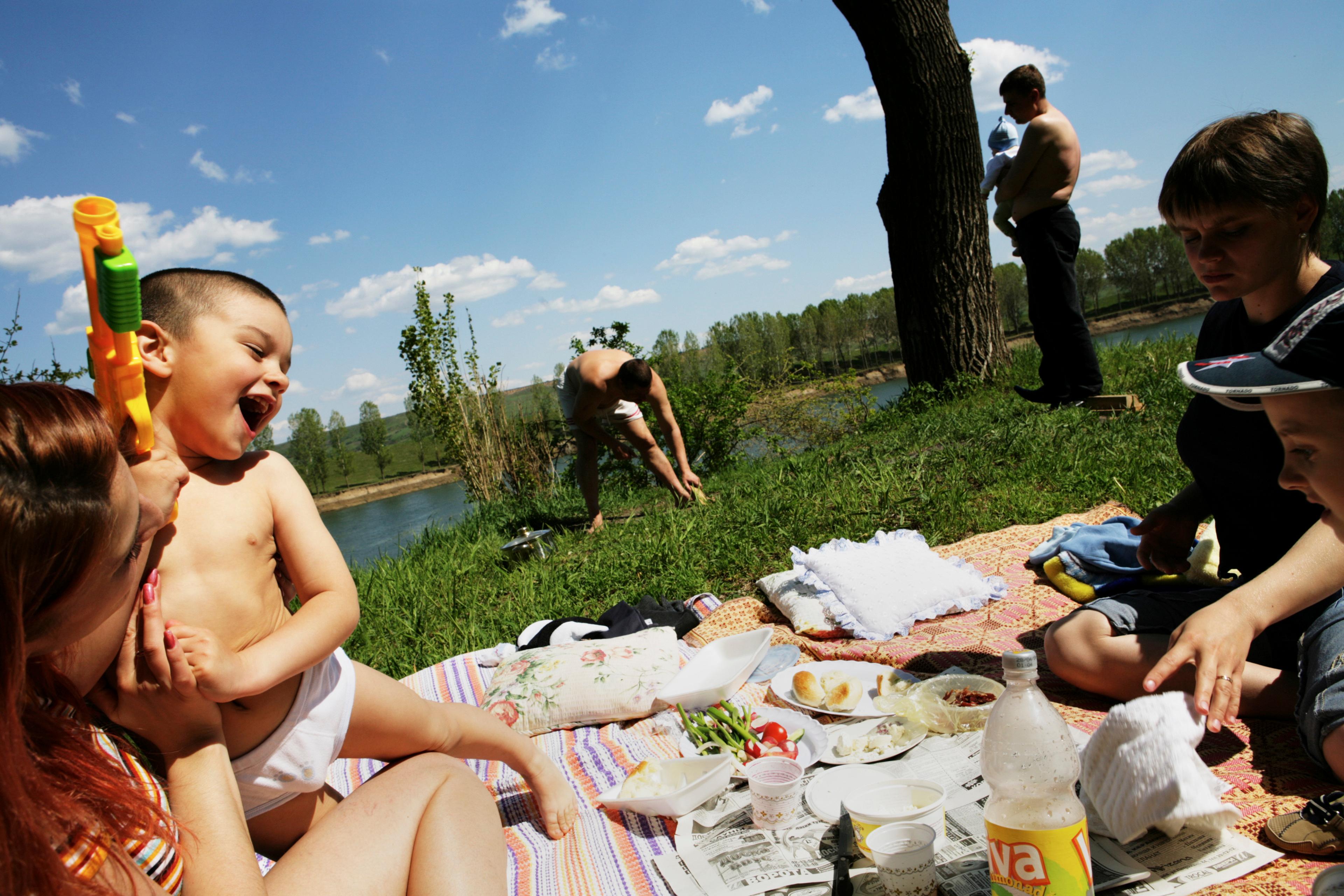A cheerful picnic by a river with adults and children relaxing on blankets under a blue sky with scattered clouds.