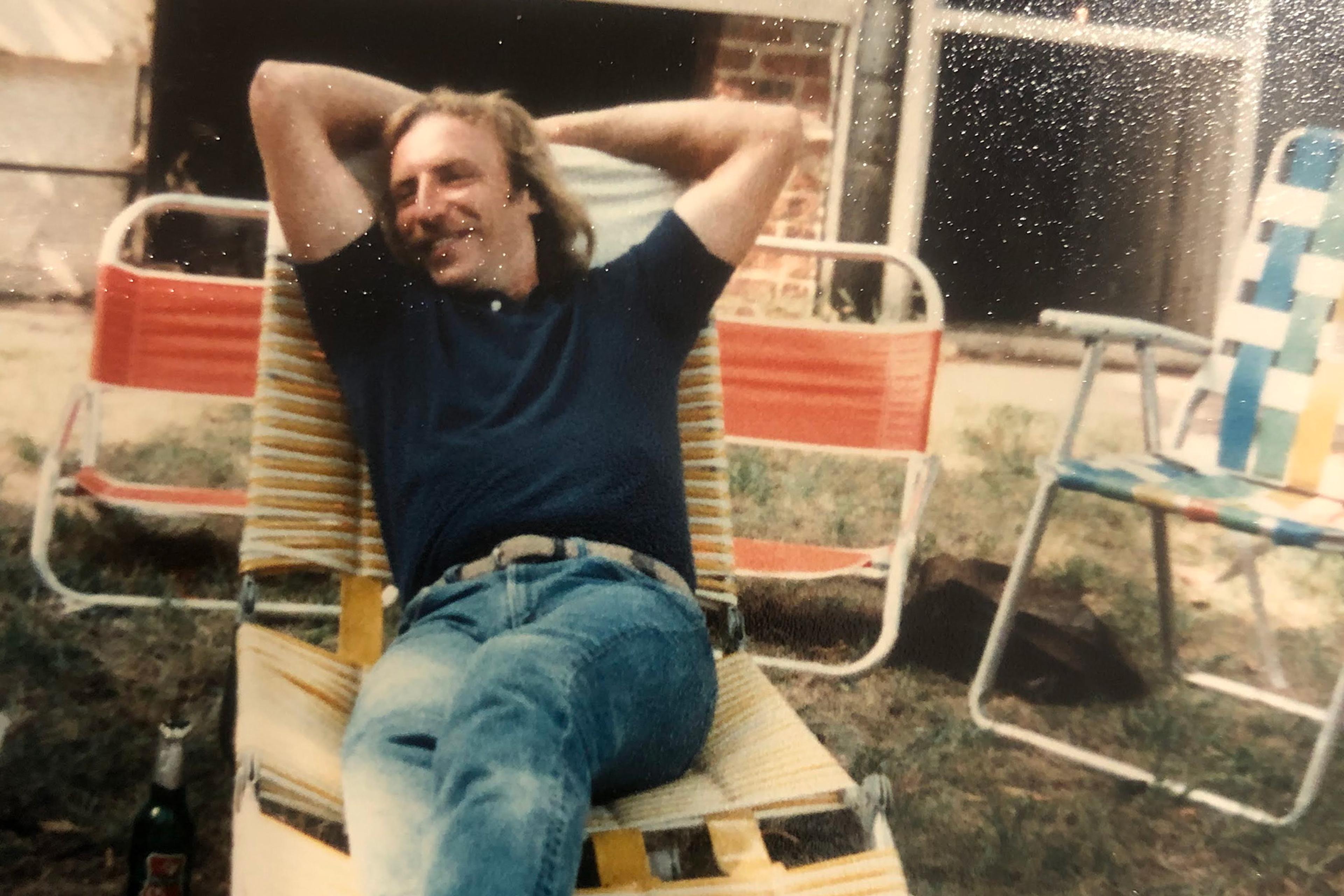 Photo of a man relaxing on a garden lounger with hands behind head smiling, surrounded by chairs and a bottle on the grass.