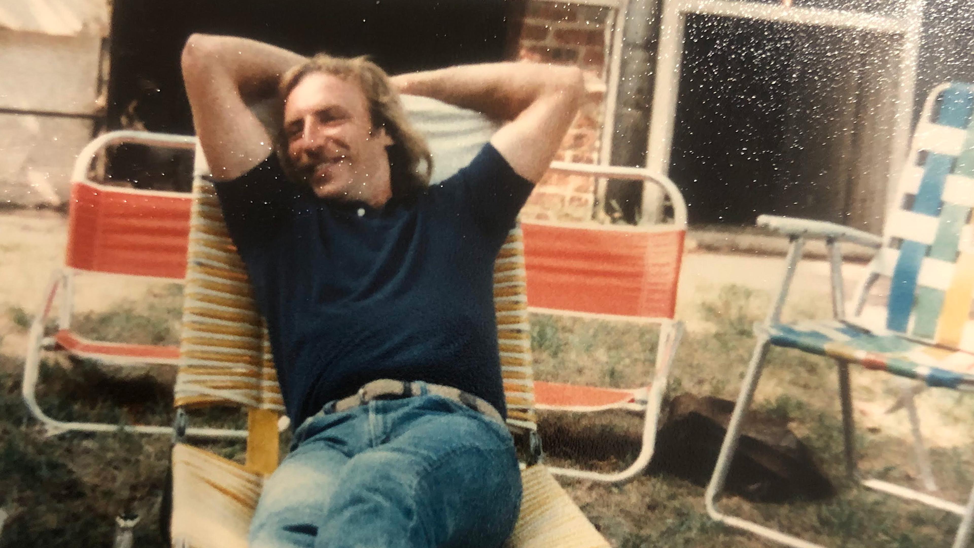 Photo of a man relaxing on a garden lounger with hands behind head smiling, surrounded by chairs and a bottle on the grass.
