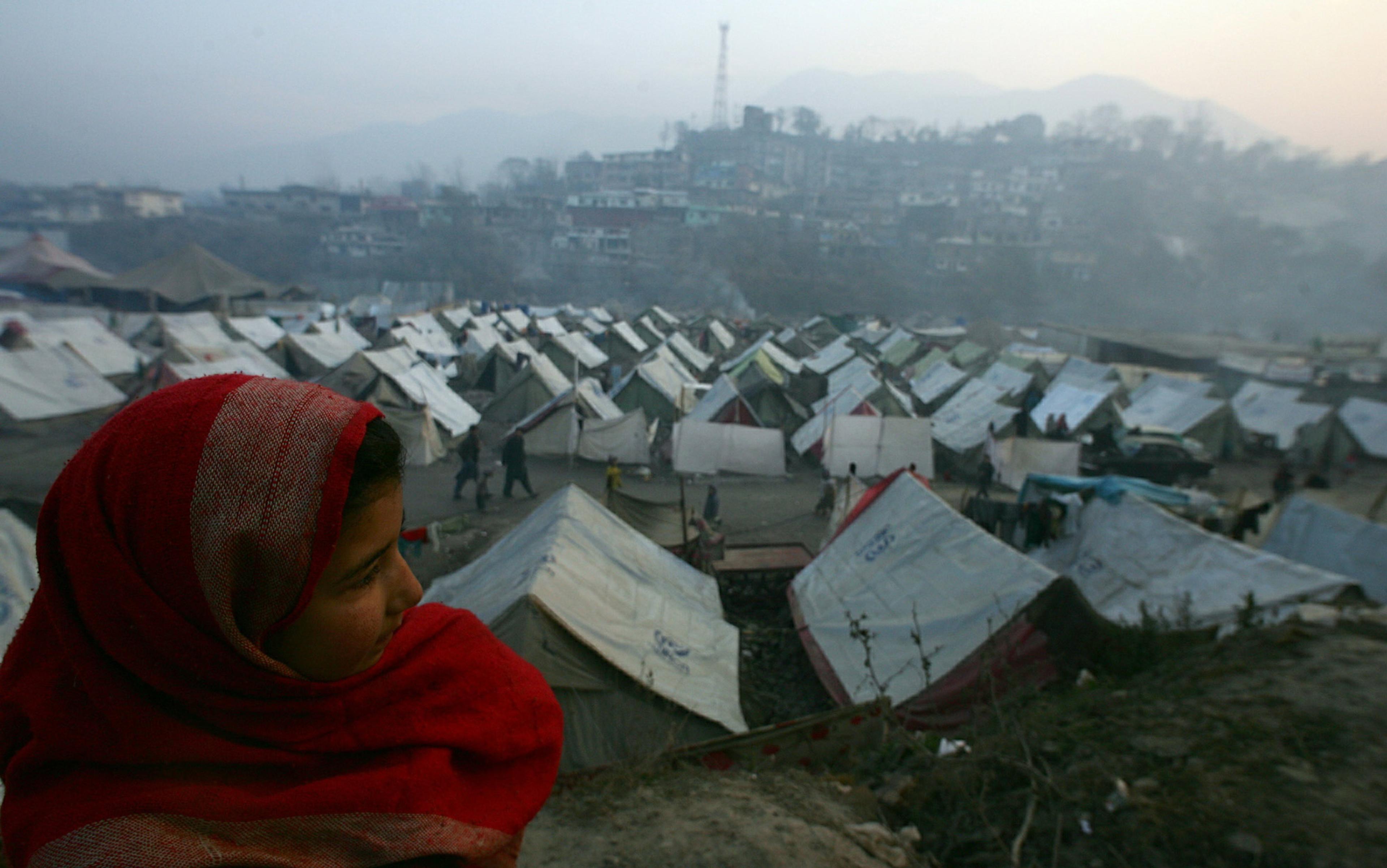 A child in a red shawl overlooking a refugee camp with numerous tents, distant buildings, and hazy mountains in the background.