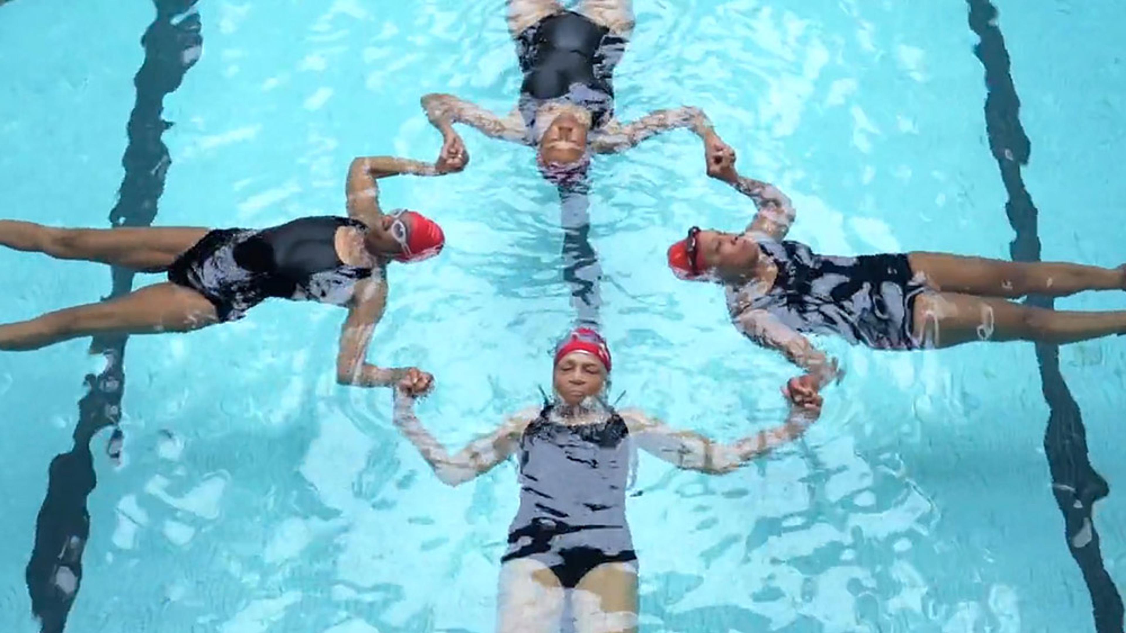 Four synchronised swimmers wearing red caps and black swimsuits holding hands, floating on their backs in a swimming pool.
