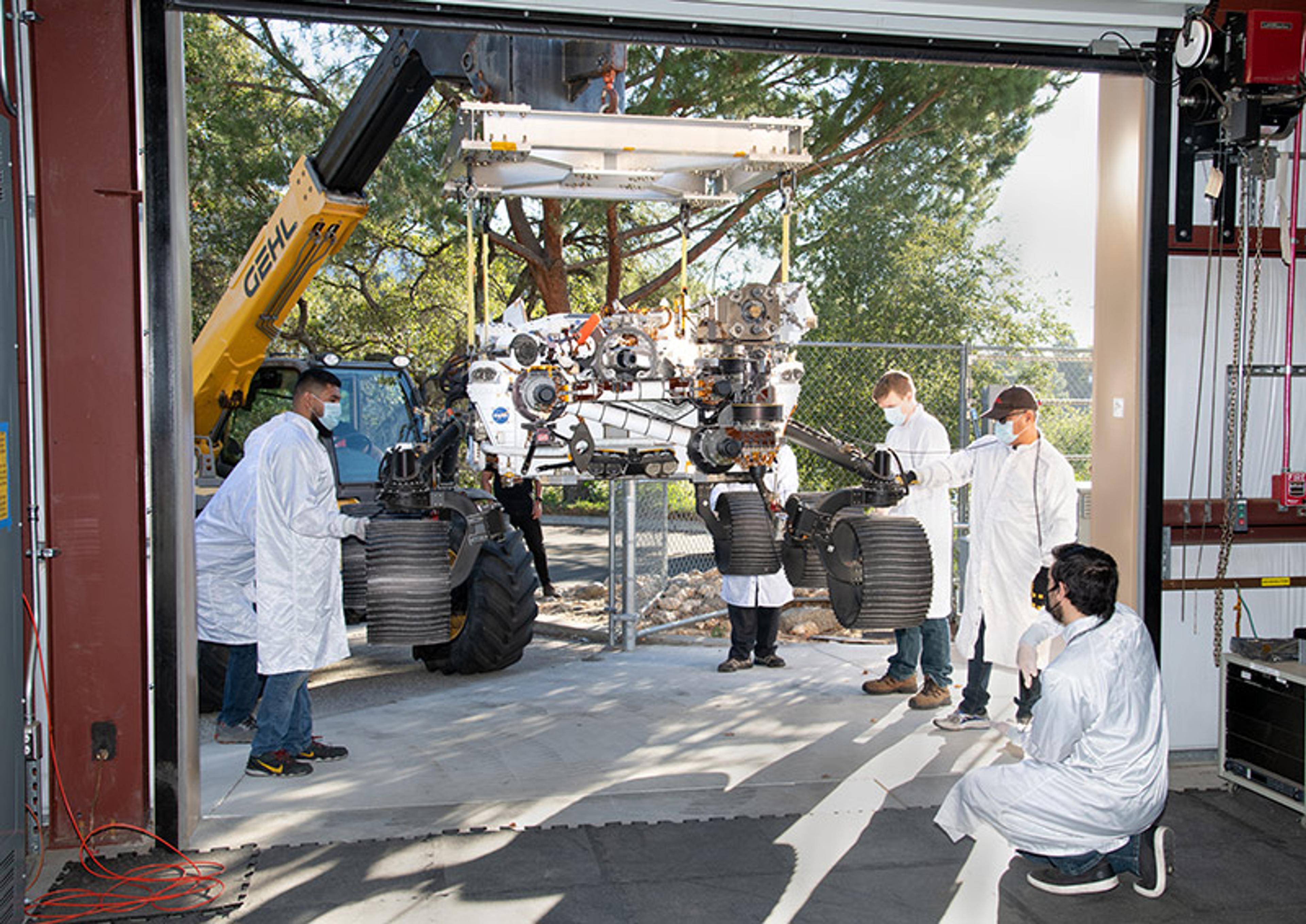 Engineers in lab coats assisting a rover being lifted by a crane, surrounded by trees and equipment in an outdoor setting.
