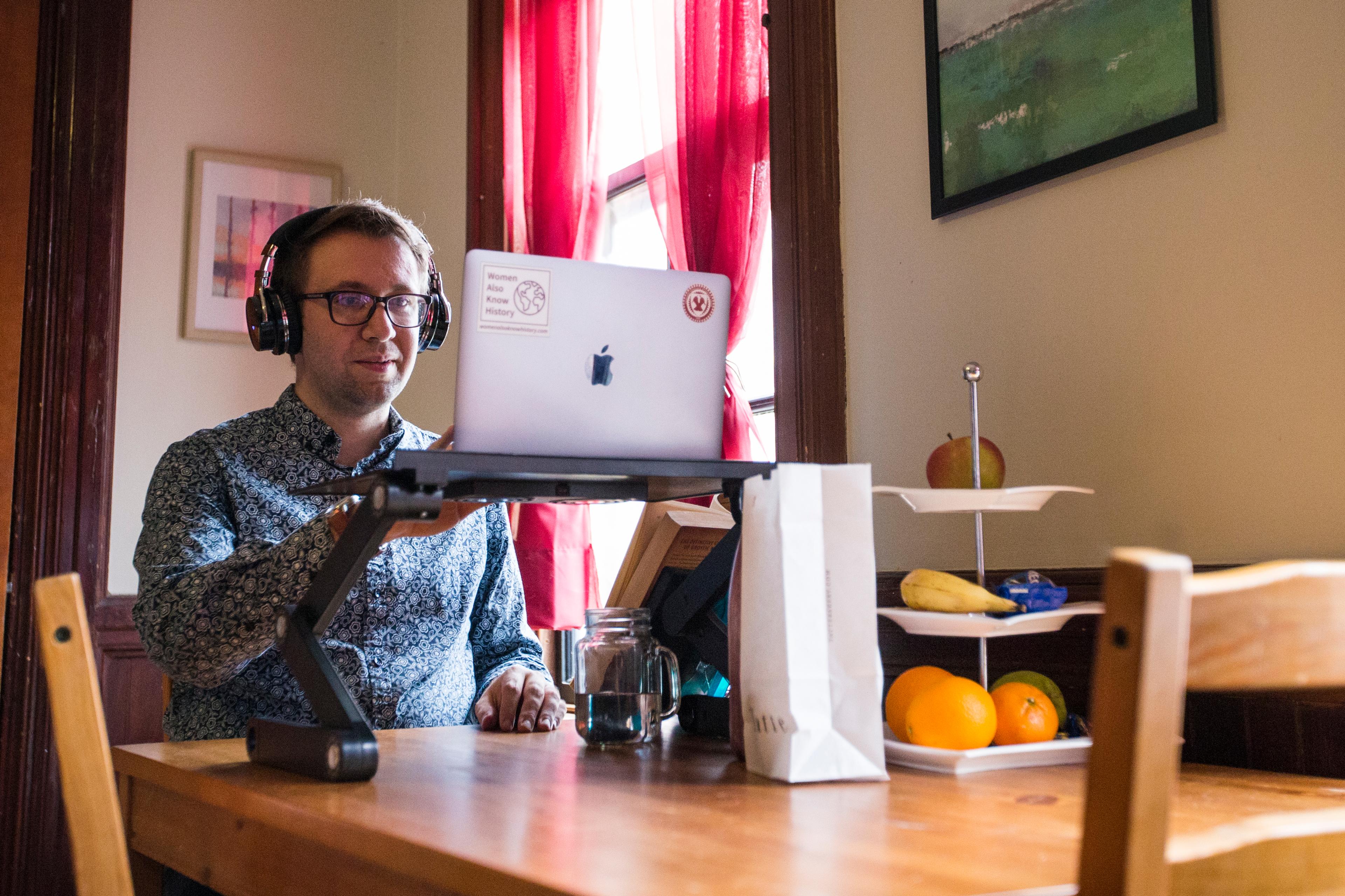 Photo of a man wearing headphones using a laptop on a stand at a wooden table with fruit on a tiered dish.