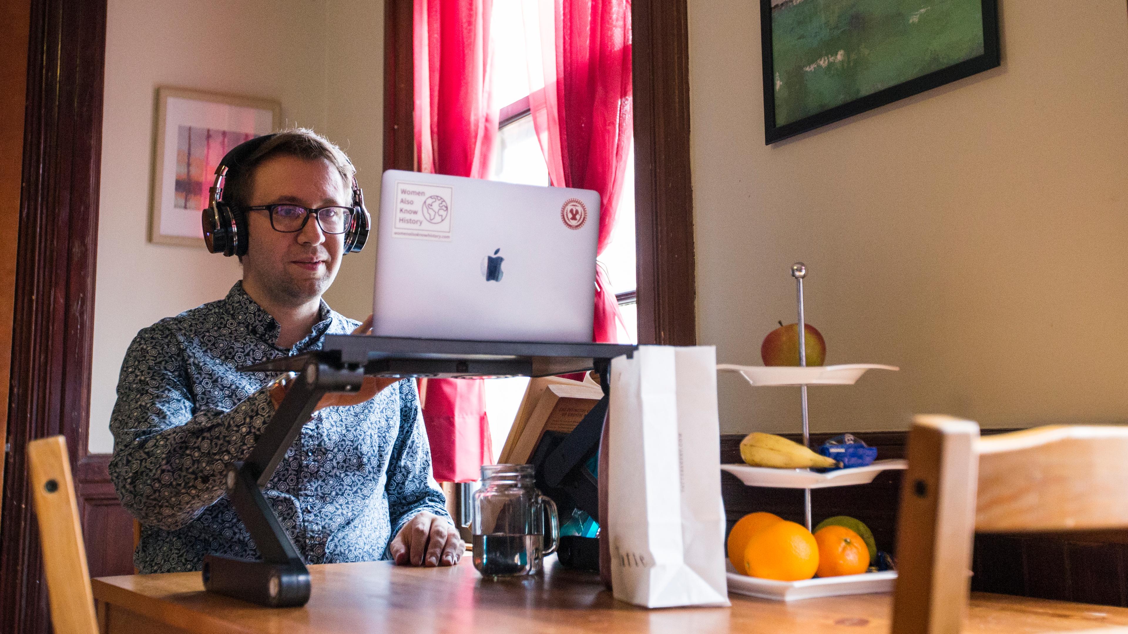Photo of a man wearing headphones using a laptop on a stand at a wooden table with fruit on a tiered dish.
