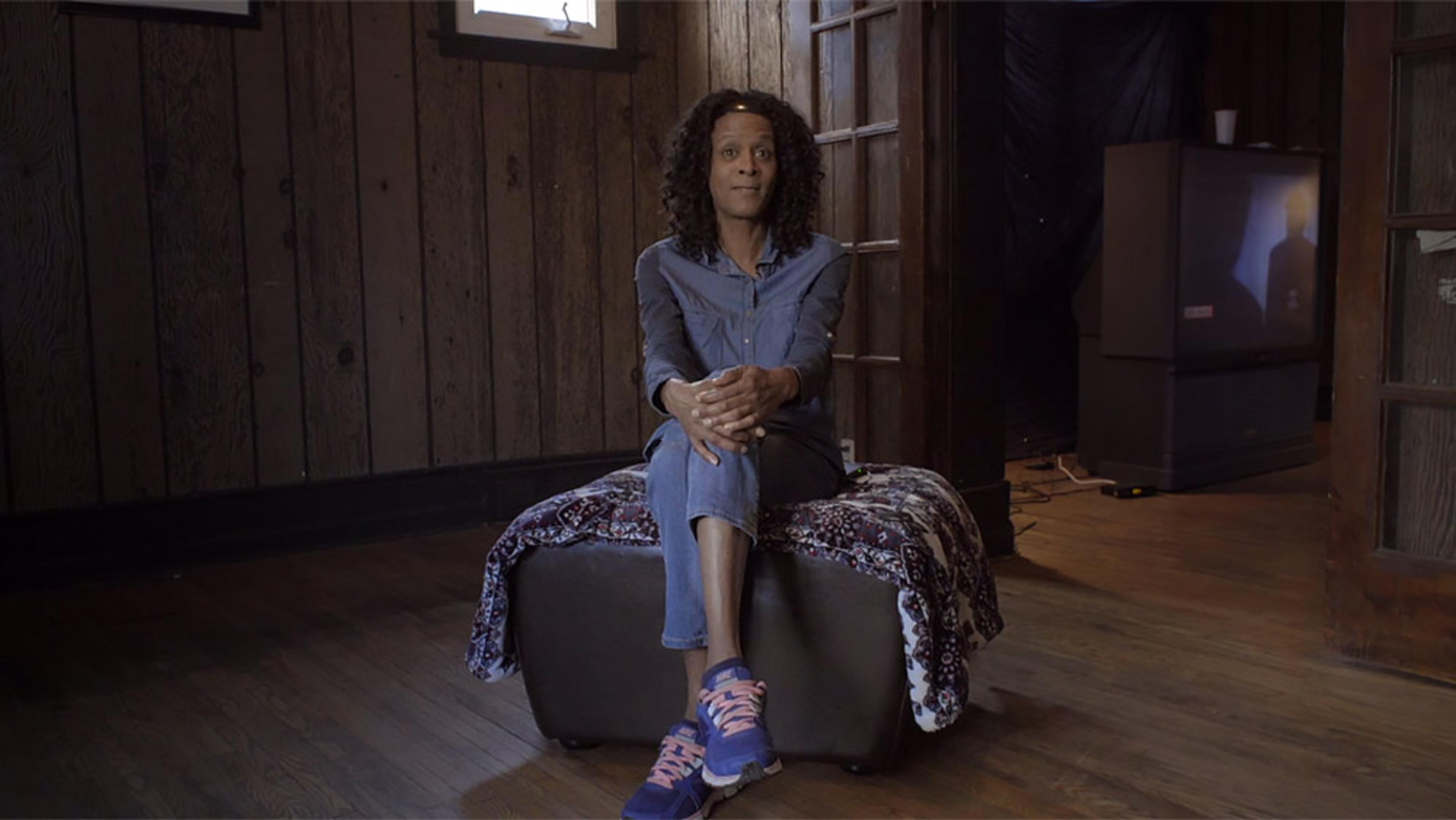 Photo of a person with curly hair sitting on a stool in a wood-panelled room, with a TV in the background to the right.