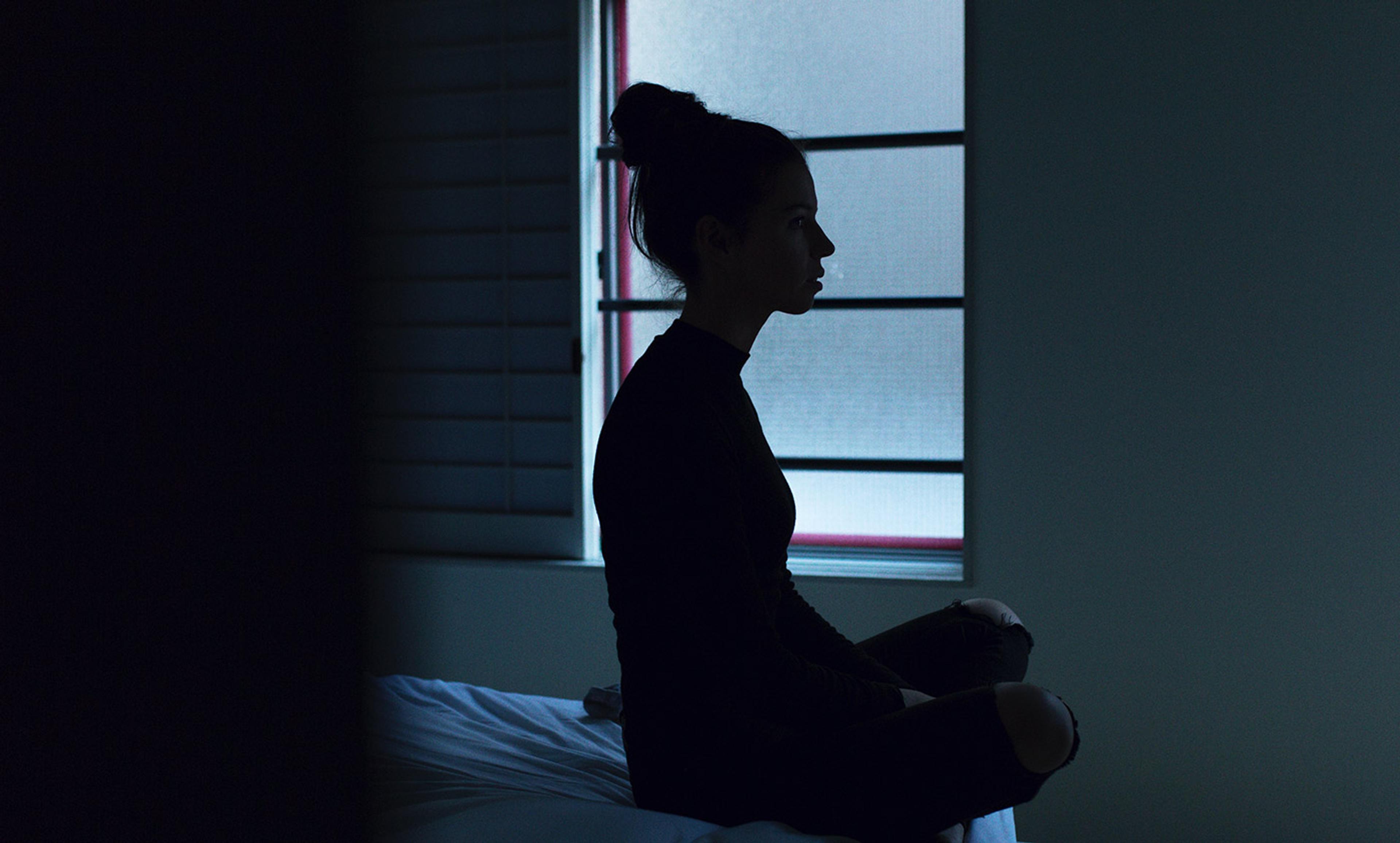 A woman in silhouette sitting crosslegged on a bed in a dimly lit room with shutters and a window in the background.