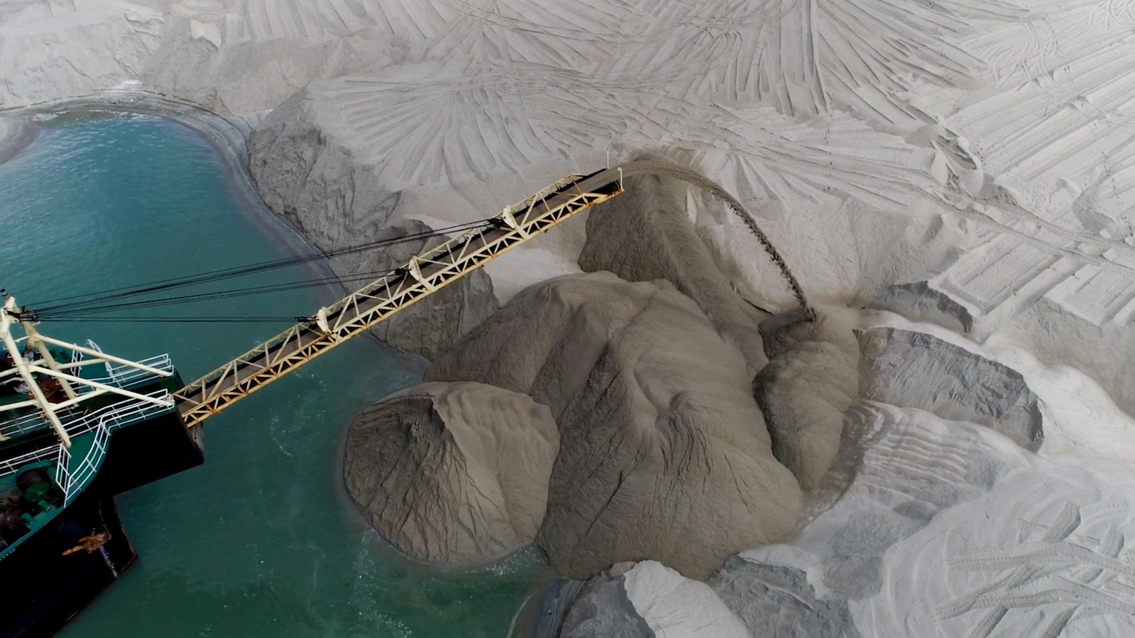 Aerial photo of an industrial conveyor unloading material into a turquoise water body, surrounded by carved sand dunes.