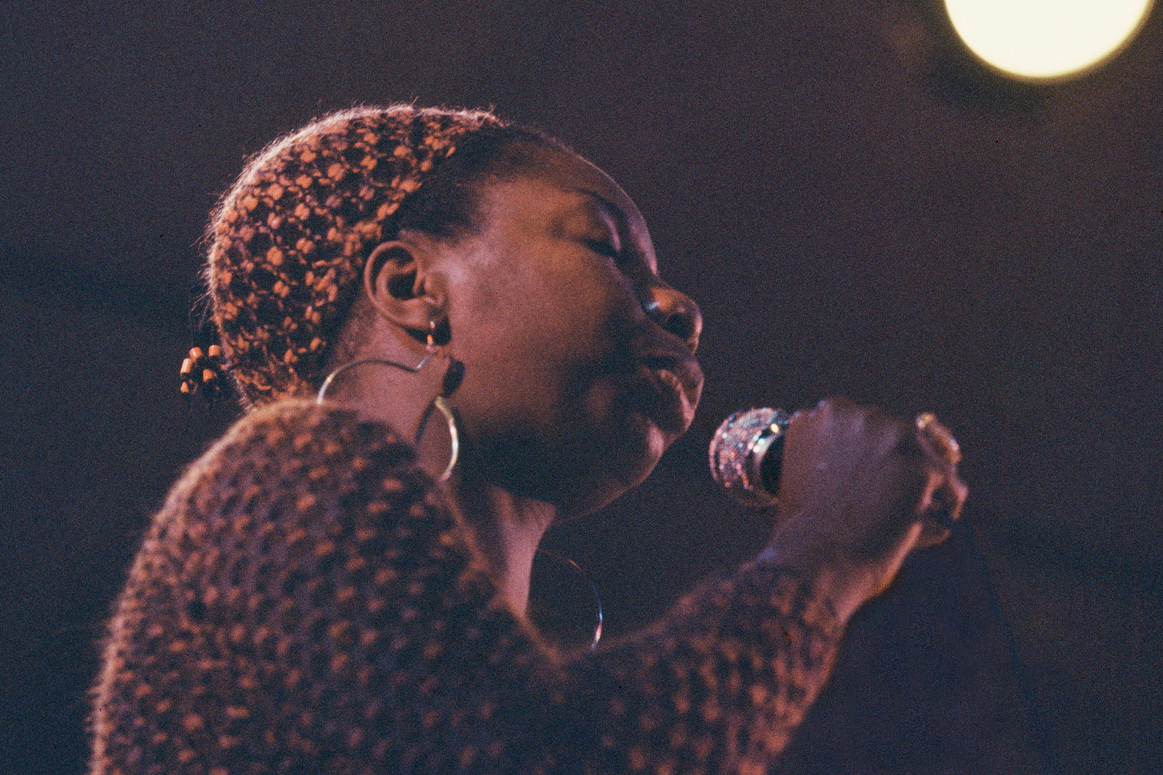 Photo of a person singing into a microphone on stage, wearing hoop earrings and a textured top, with a spotlight in the background.