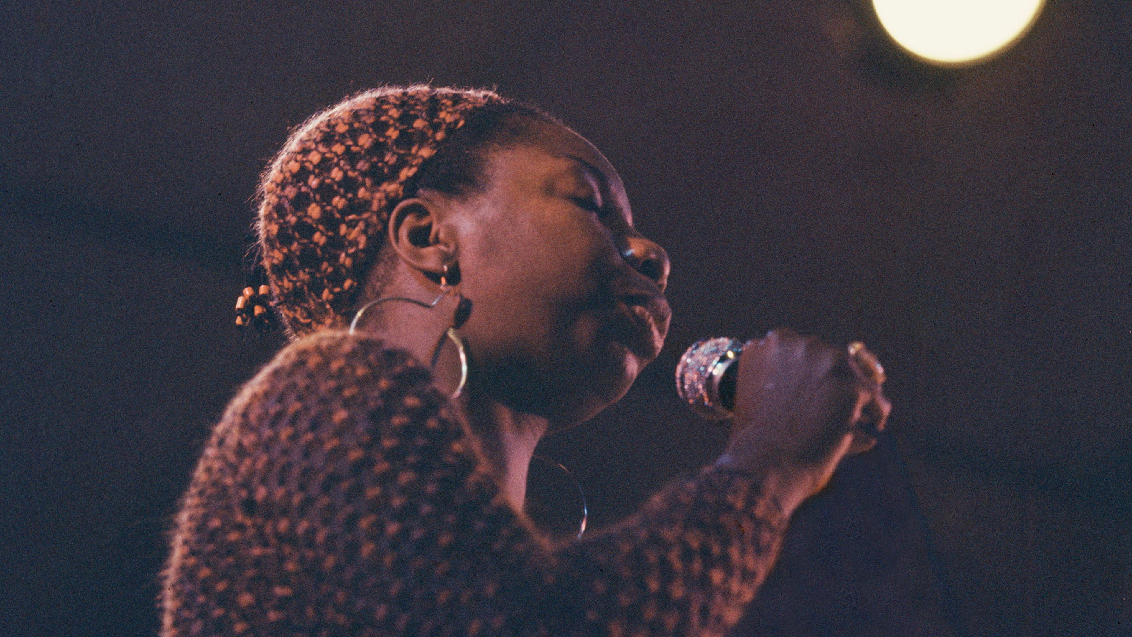 Photo of a person singing into a microphone on stage, wearing hoop earrings and a textured top, with a spotlight in the background.