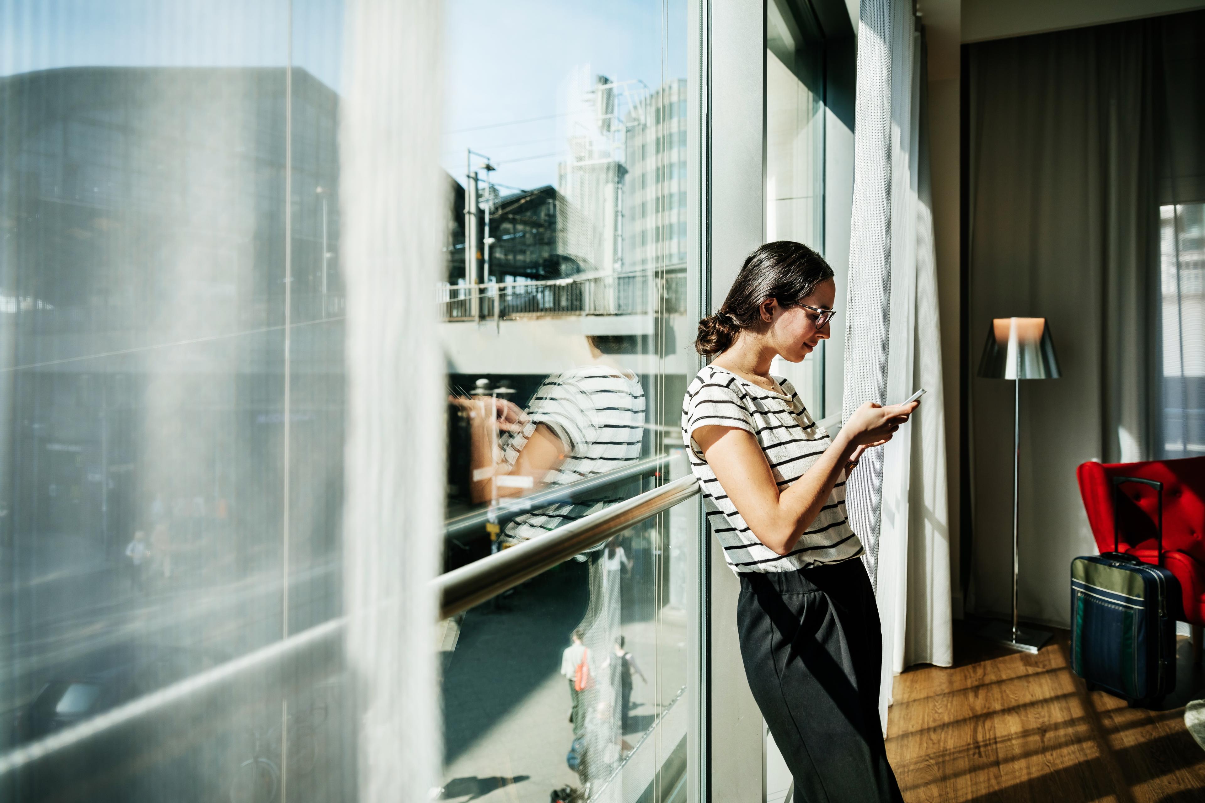 Photo of a woman using a phone by a sunny window with a city view. She is next to a suitcase and a red chair.
