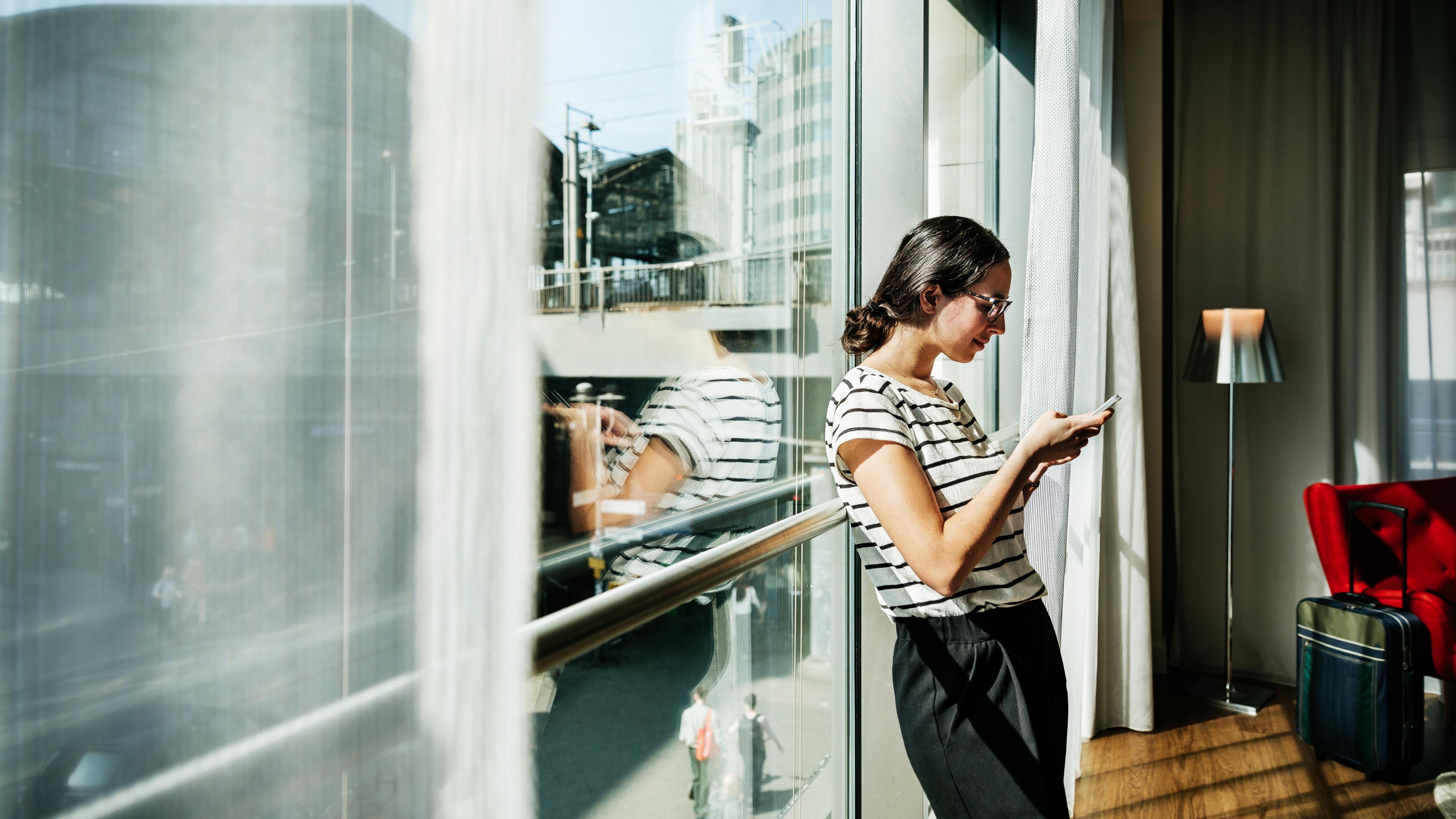 Photo of a woman using a phone by a sunny window with a city view. She is next to a suitcase and a red chair.