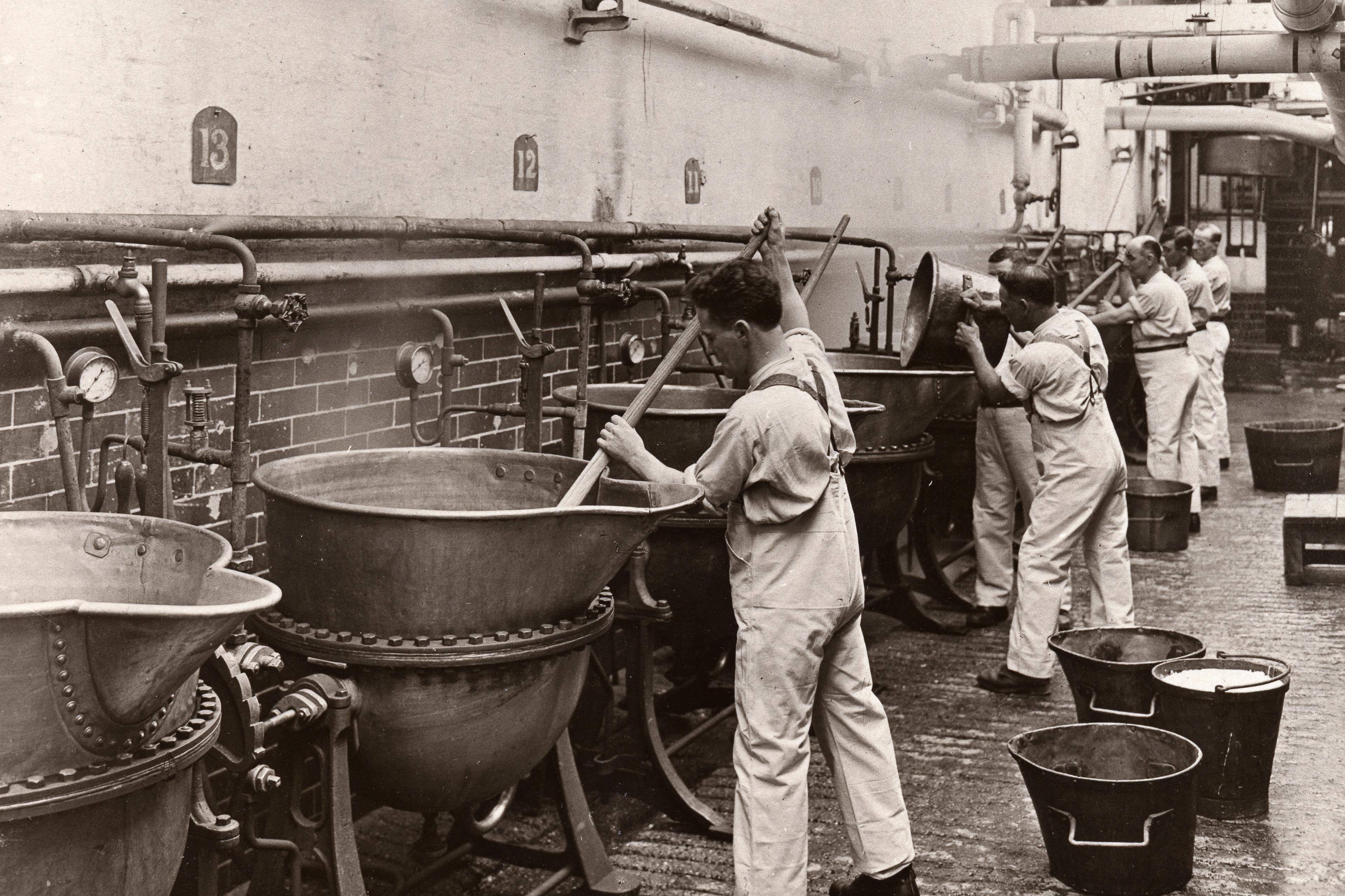 Black and white photo of workers in a factory stirring large metal vats with wooden paddles.
