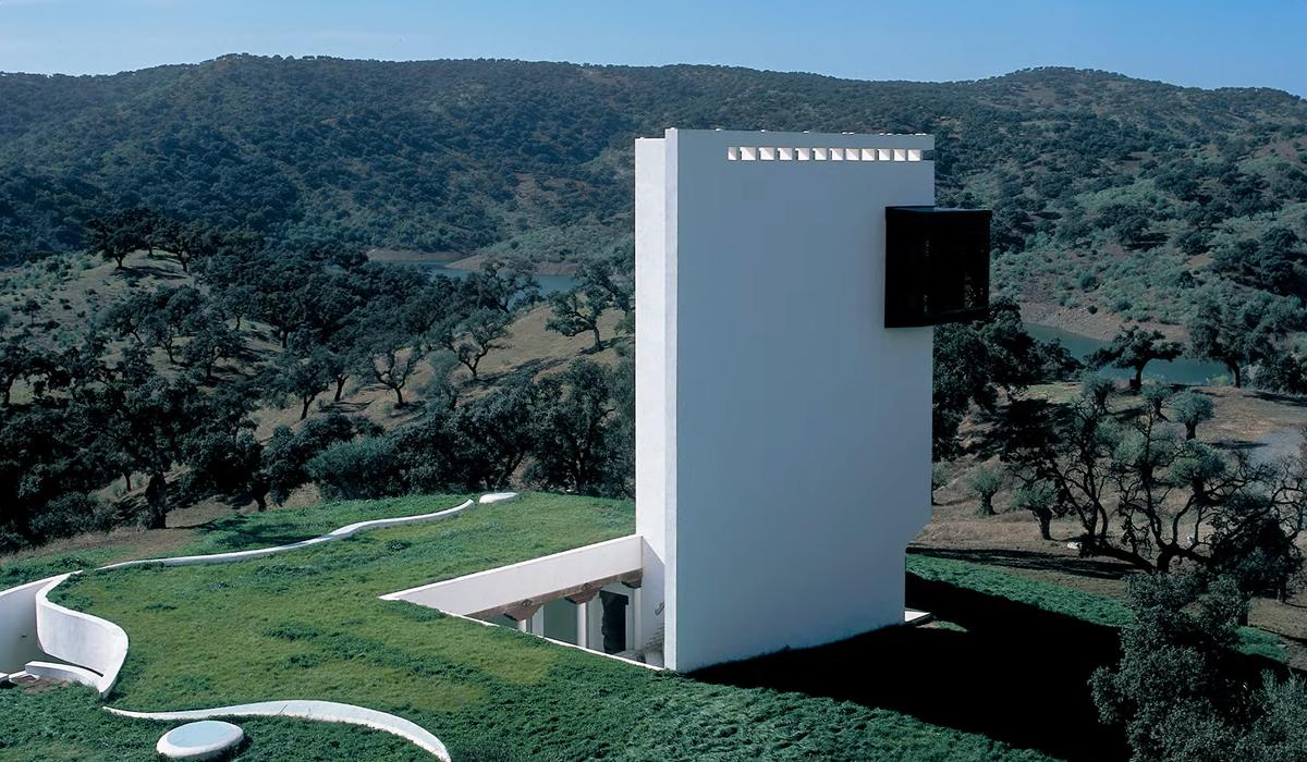 Modern white tower building amidst green landscape with winding path and trees, hills and a clear sky in the background.