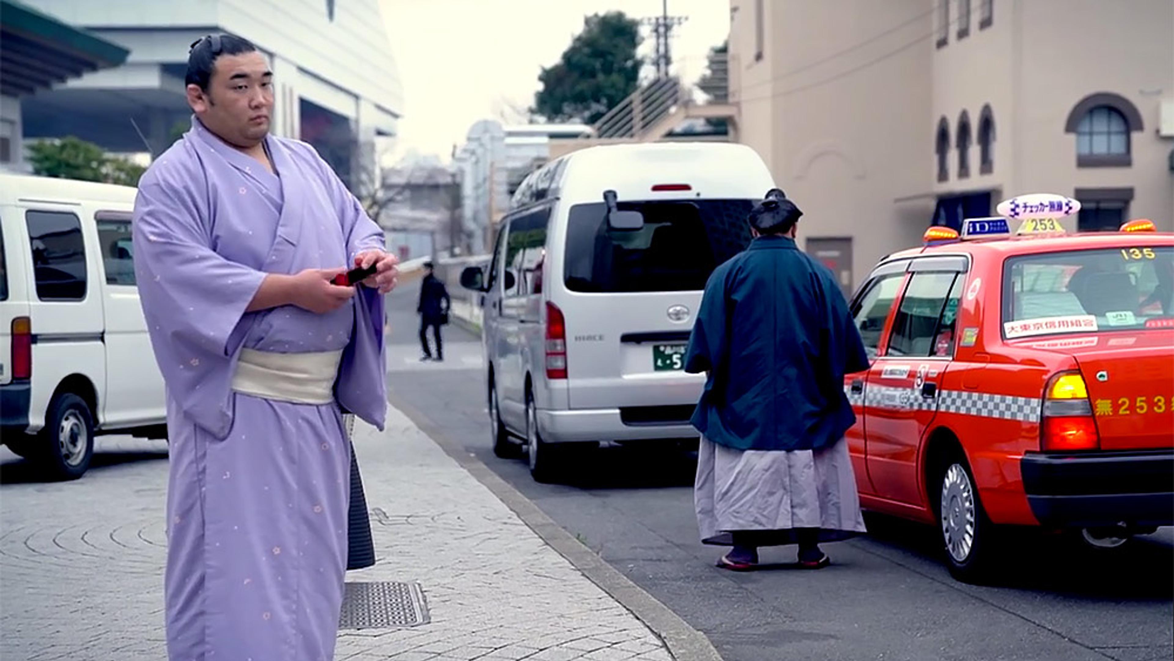 A Japanese man in a lavender kimono on a street with vans and a red taxi, another man in traditional attire near the taxi.