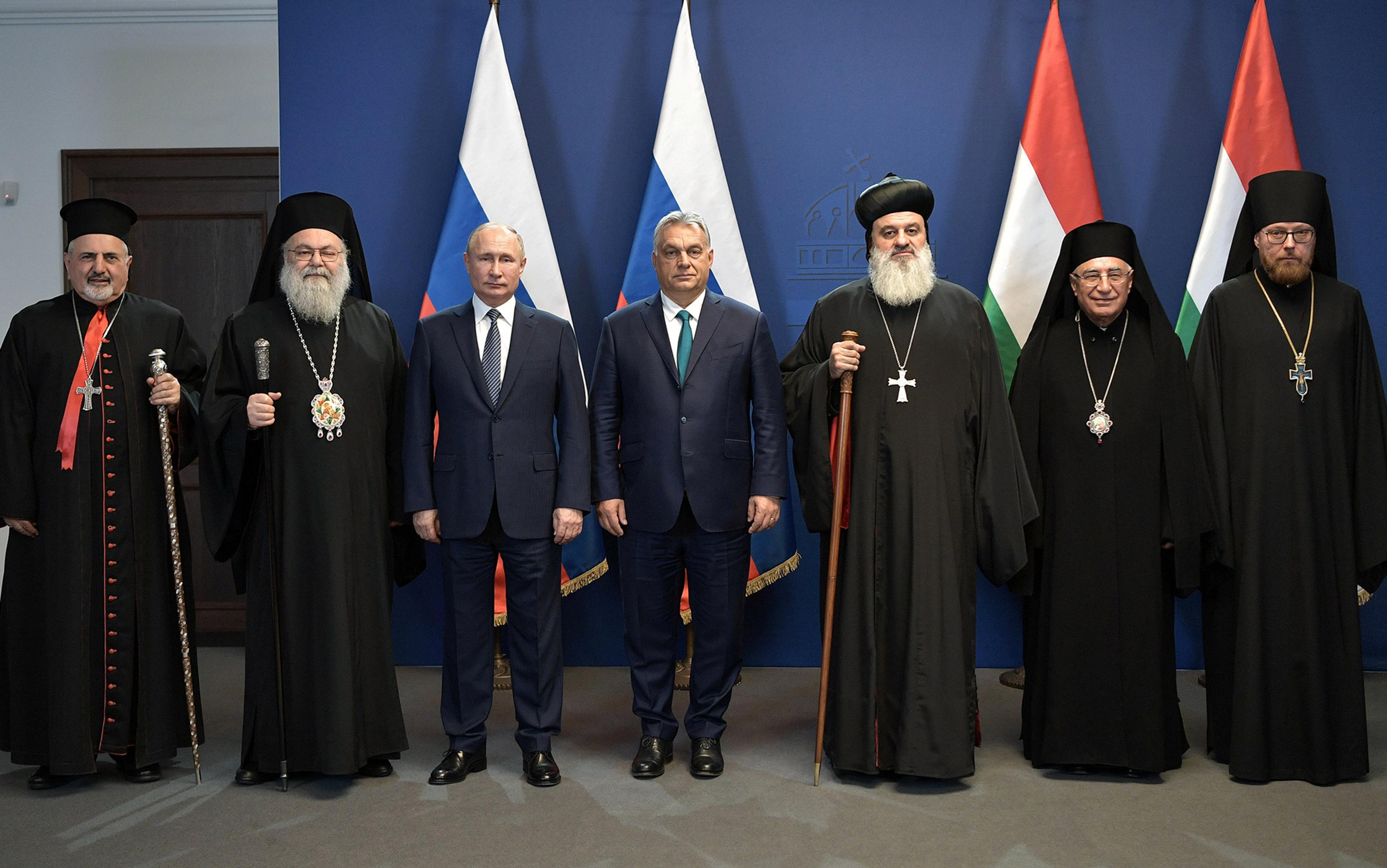 A group of men, including religious leaders in robes and two suited individuals, standing in front of Russian and Hungarian flags.