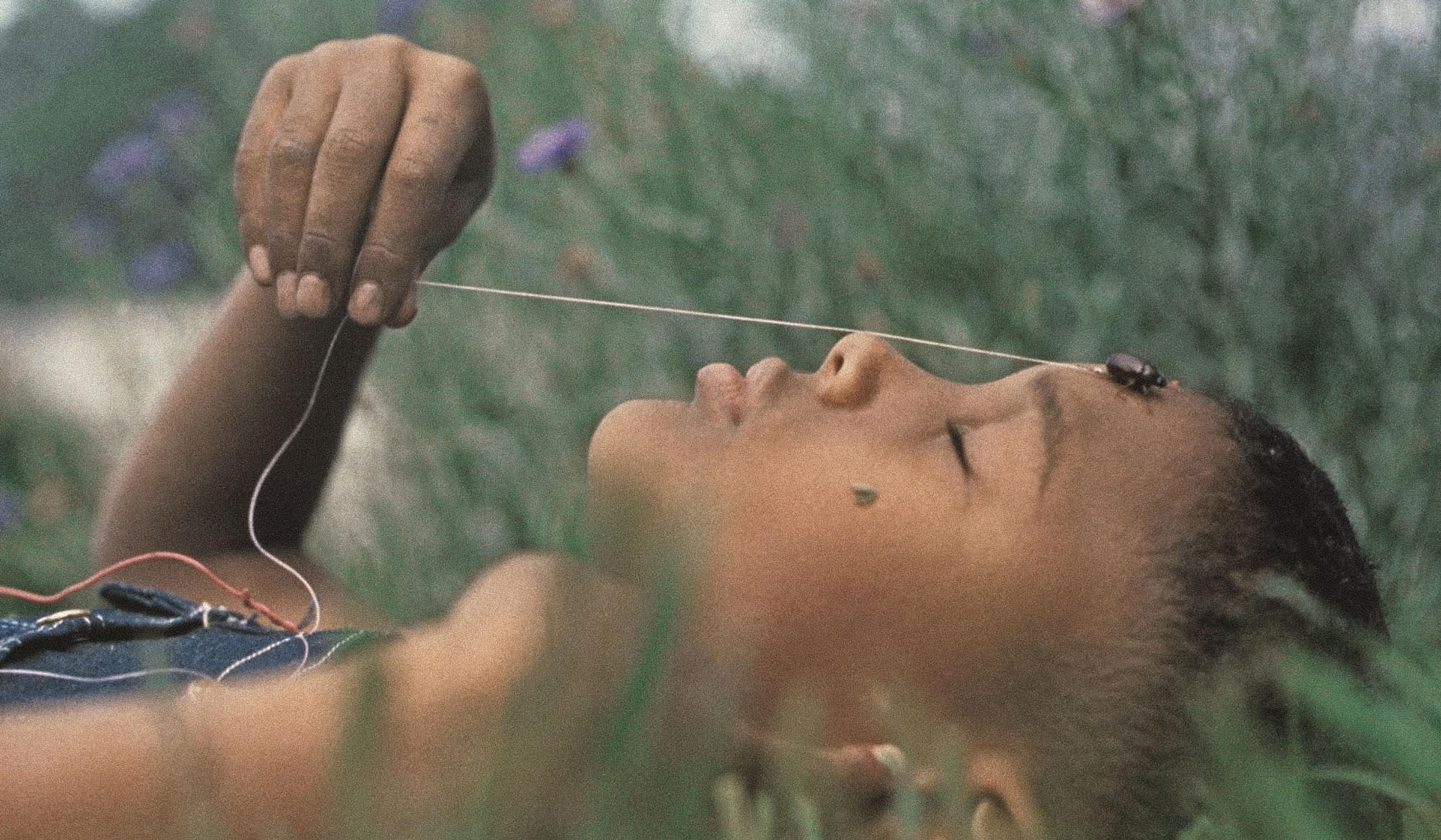 Close-up of a child lying in grass with eyes closed, holding a string tied to a beetle on their forehead, surrounded by greenery.