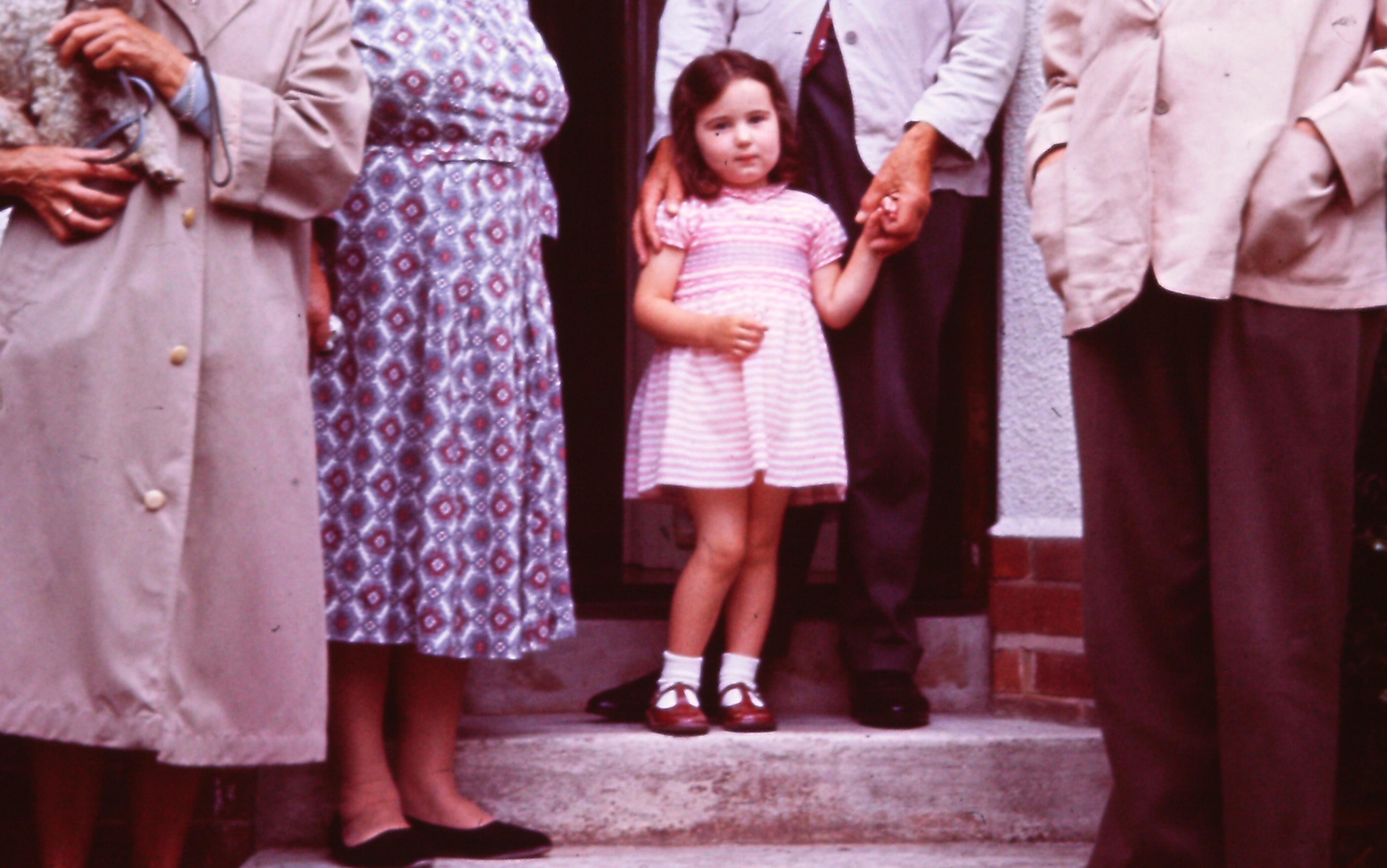 A young girl in a pink dress stands on a step, holding the hand of an adult. Four adults are partially visible around her.