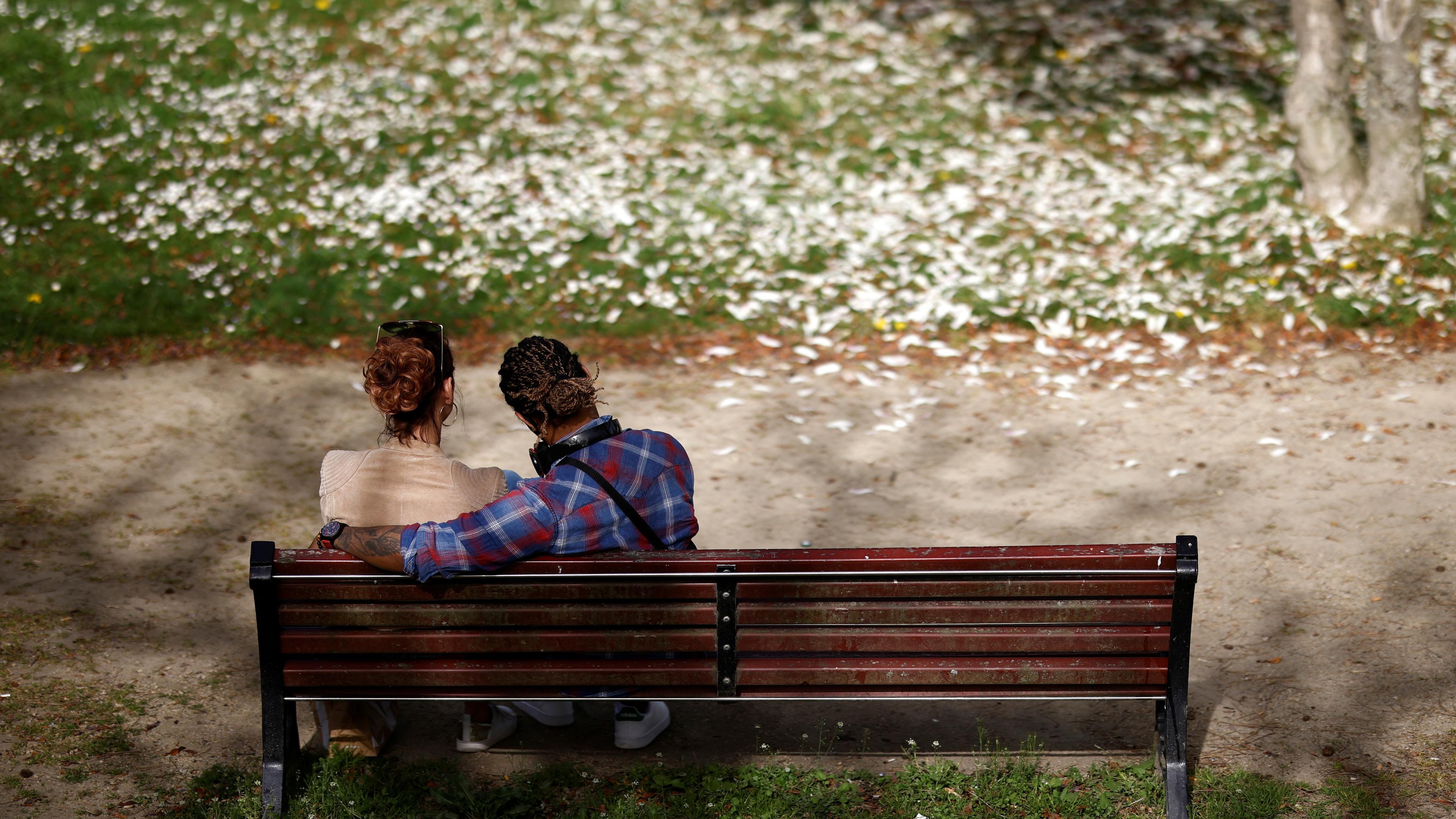 Photo of two people sitting on a park bench, one with their arm around the other, surrounded by grass and fallen petals.