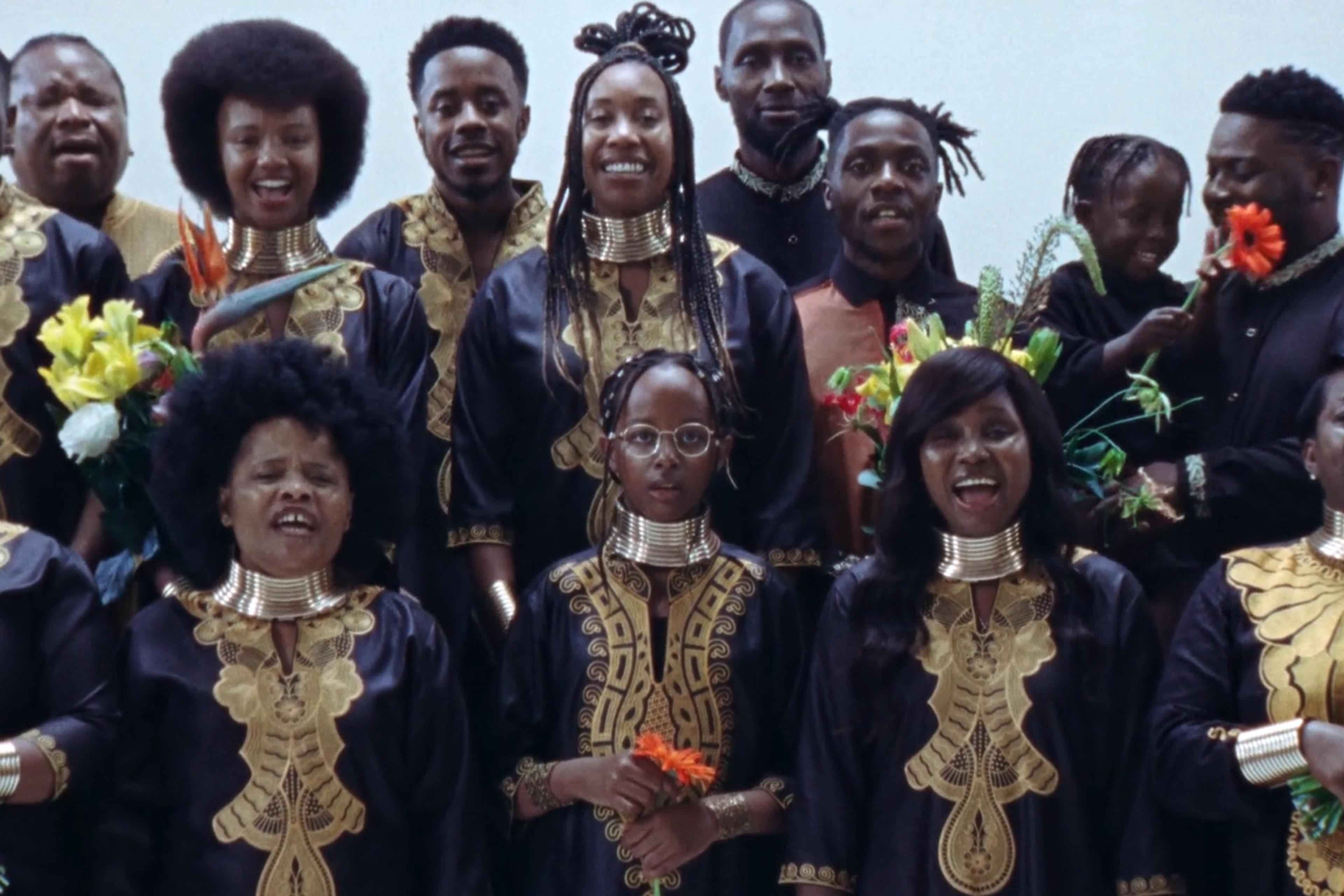 A group of people in traditional attire with gold embroidery, holding flowers and smiling while singing.